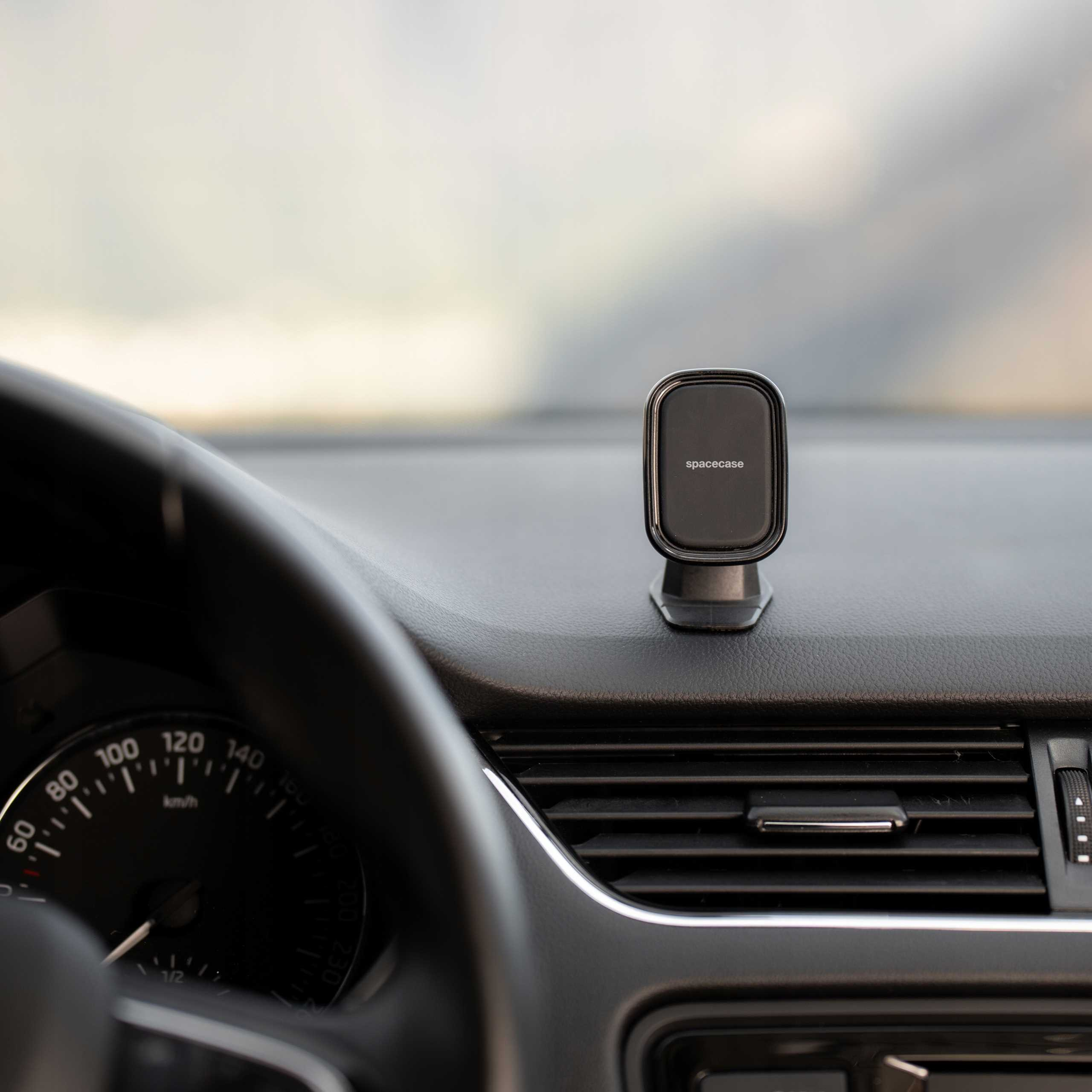 Wnętrze samochodu: deska rozdzielcza, kierownica, prędkościomierz, uchwyt na telefon z logo "spacecase". Tło rozmyte.