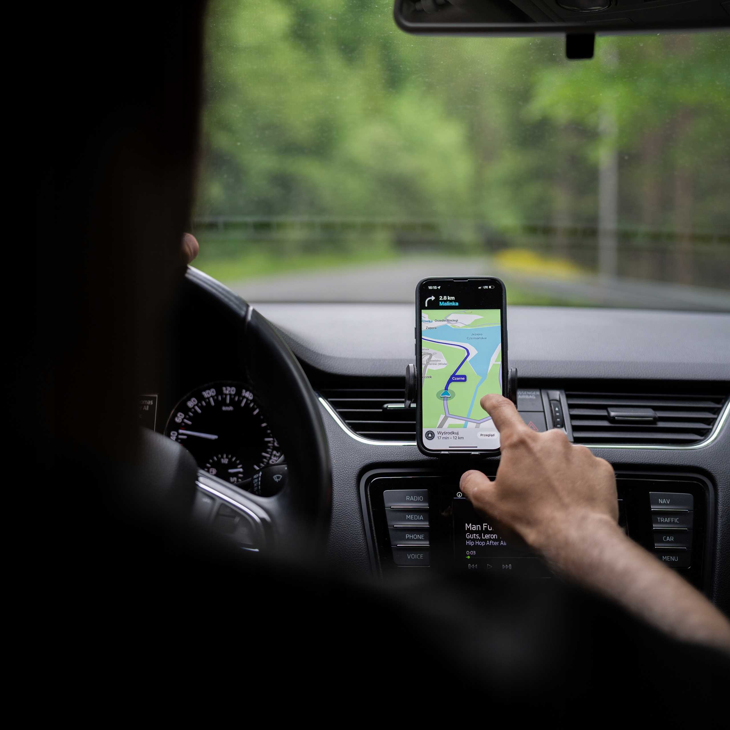 Osoba prowadzi auto, używając telefonu z nawigacją. Telefon pokazuje mapę i jest zamontowany na desce rozdzielczej.