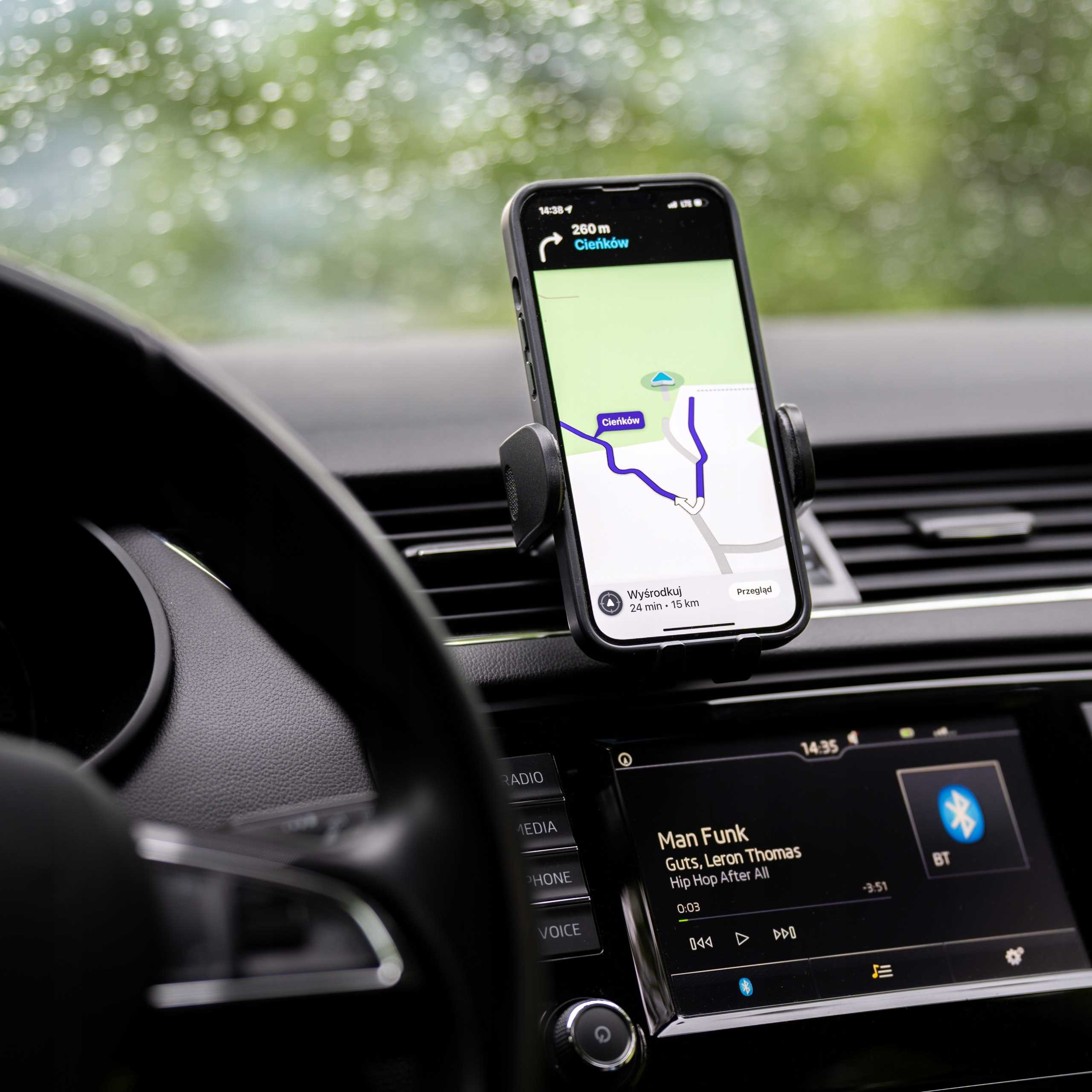 Wnętrze samochodu z telefonem pokazującym nawigację, kierownicą i ekranem informacyjnym. Zielone tło przez przednią szybę.