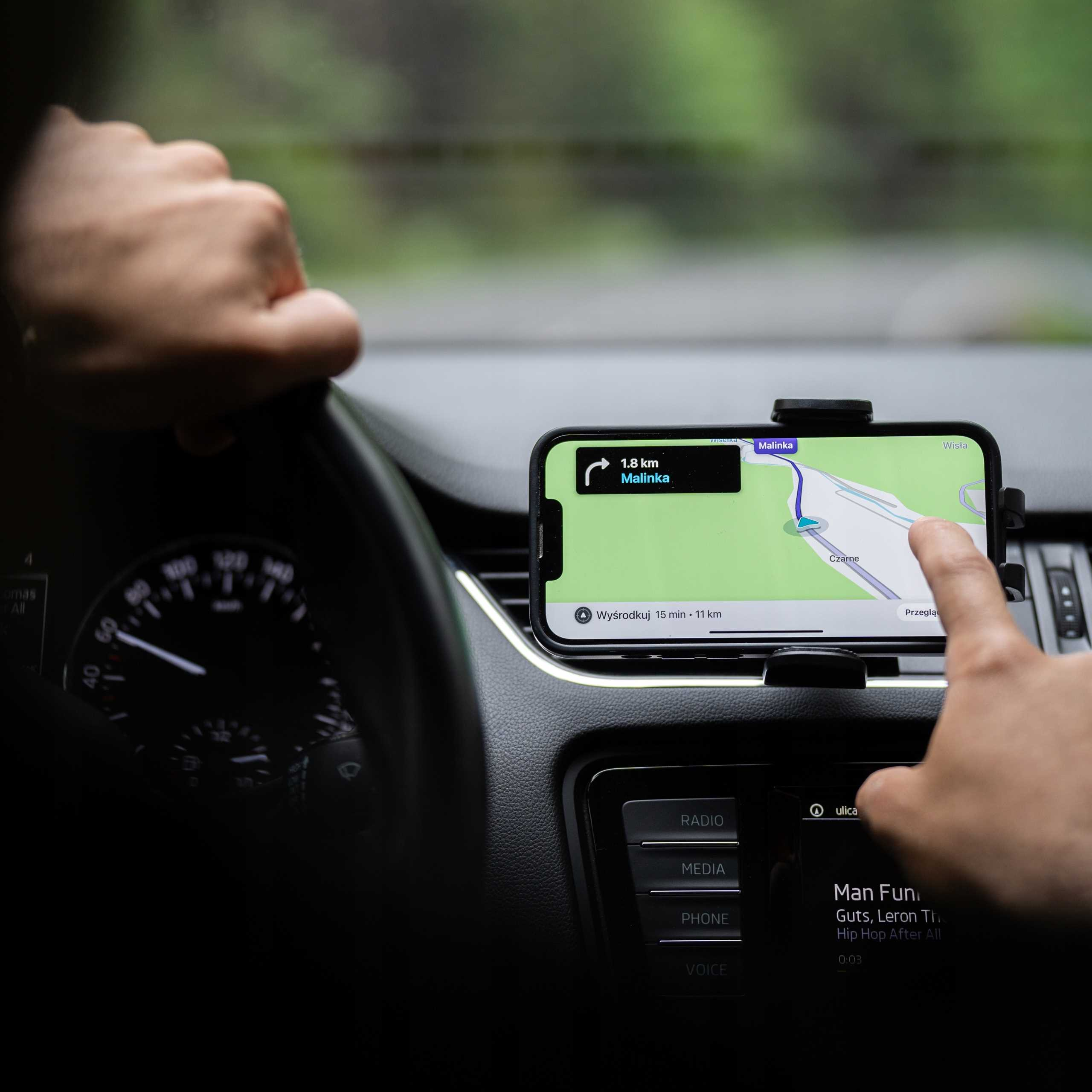 Ręka dotyka telefonu z mapą pokazującą wskazówki. Druga ręka na kierownicy. Wnętrze samochodu, deska rozdzielcza i tarcza widoczne.