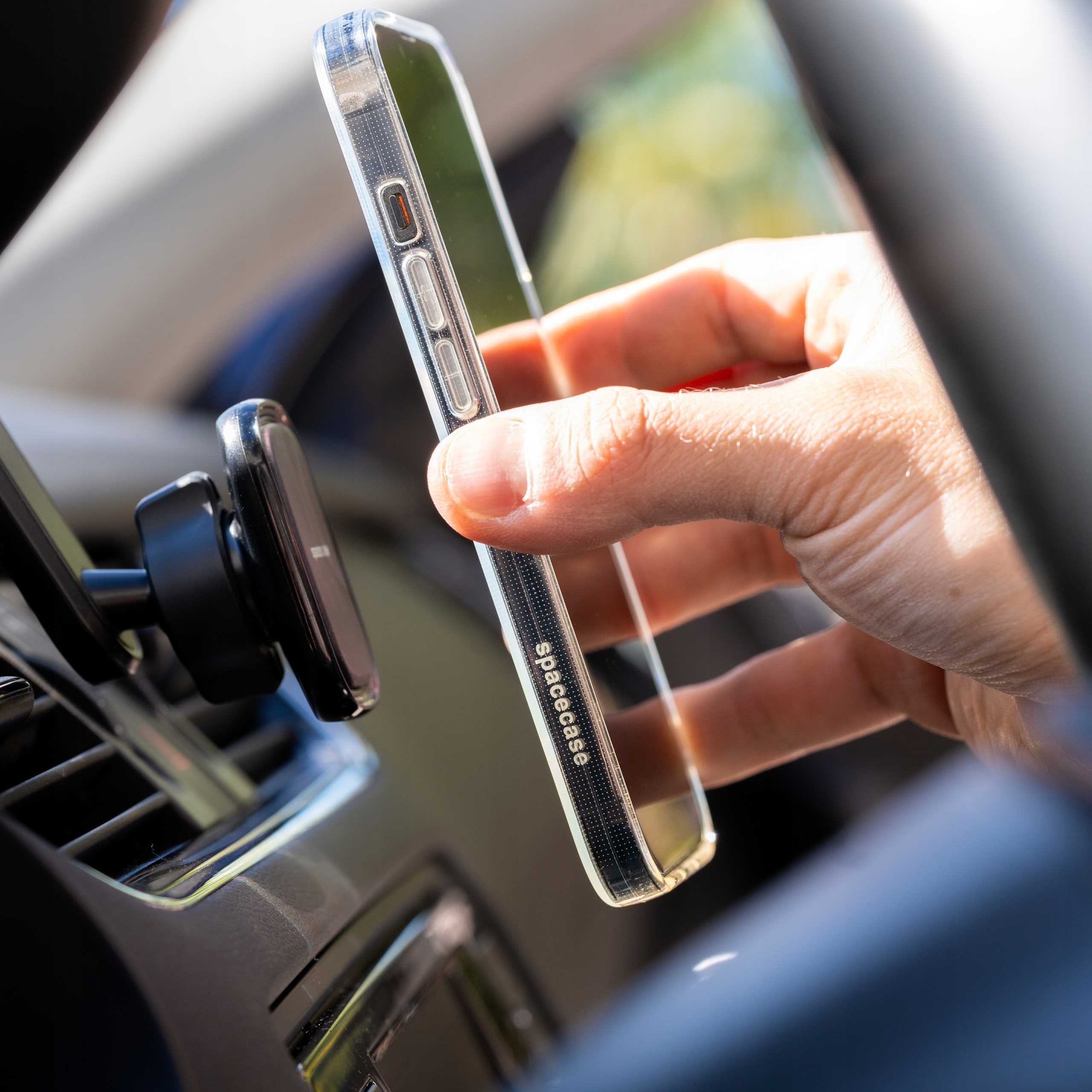 Ręka trzymająca telefon w samochodzie. Telefon jest w przezroczystym etui i zamontowany na kratce wentylacyjnej deski rozdzielczej.