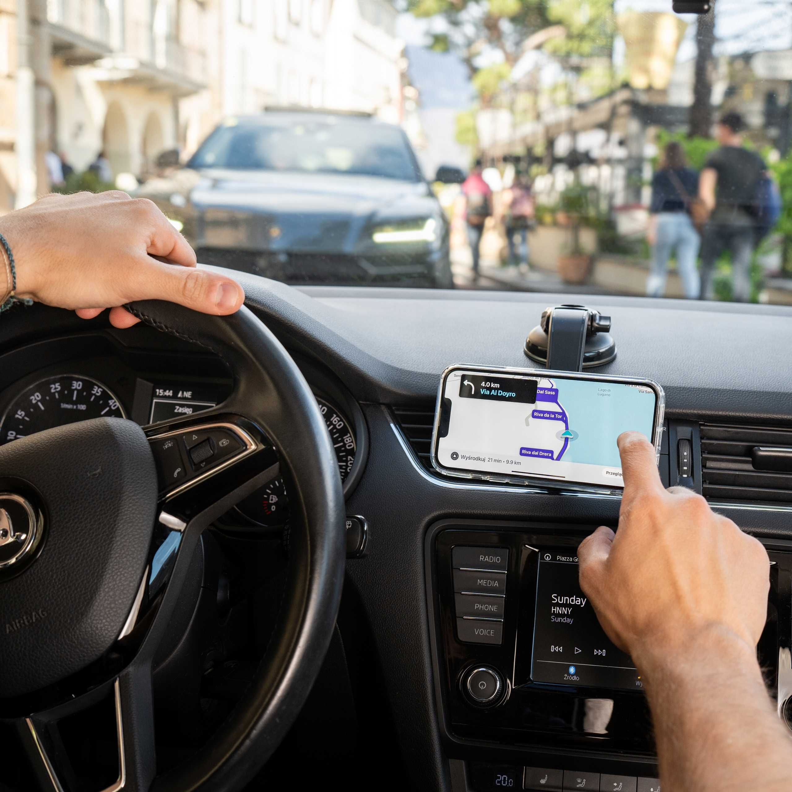 Osoba w samochodzie używa telefonu z mapą. Widoczna kierownica i deska rozdzielcza. Drugi samochód z przodu.