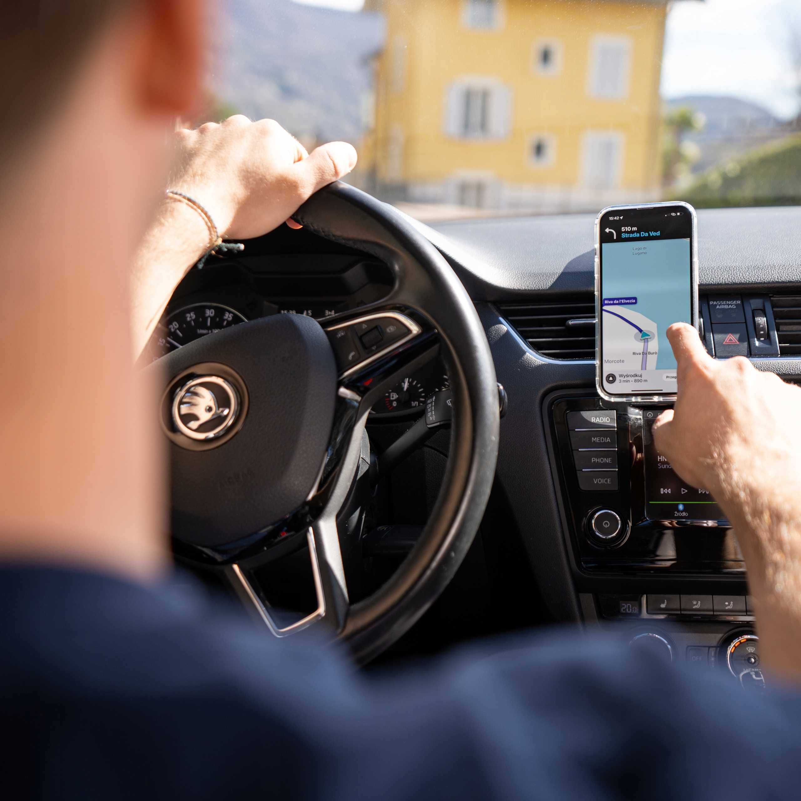 Osoba prowadzi samochód, używając telefonu do nawigacji. Przez okno widoczny jest żółty budynek.