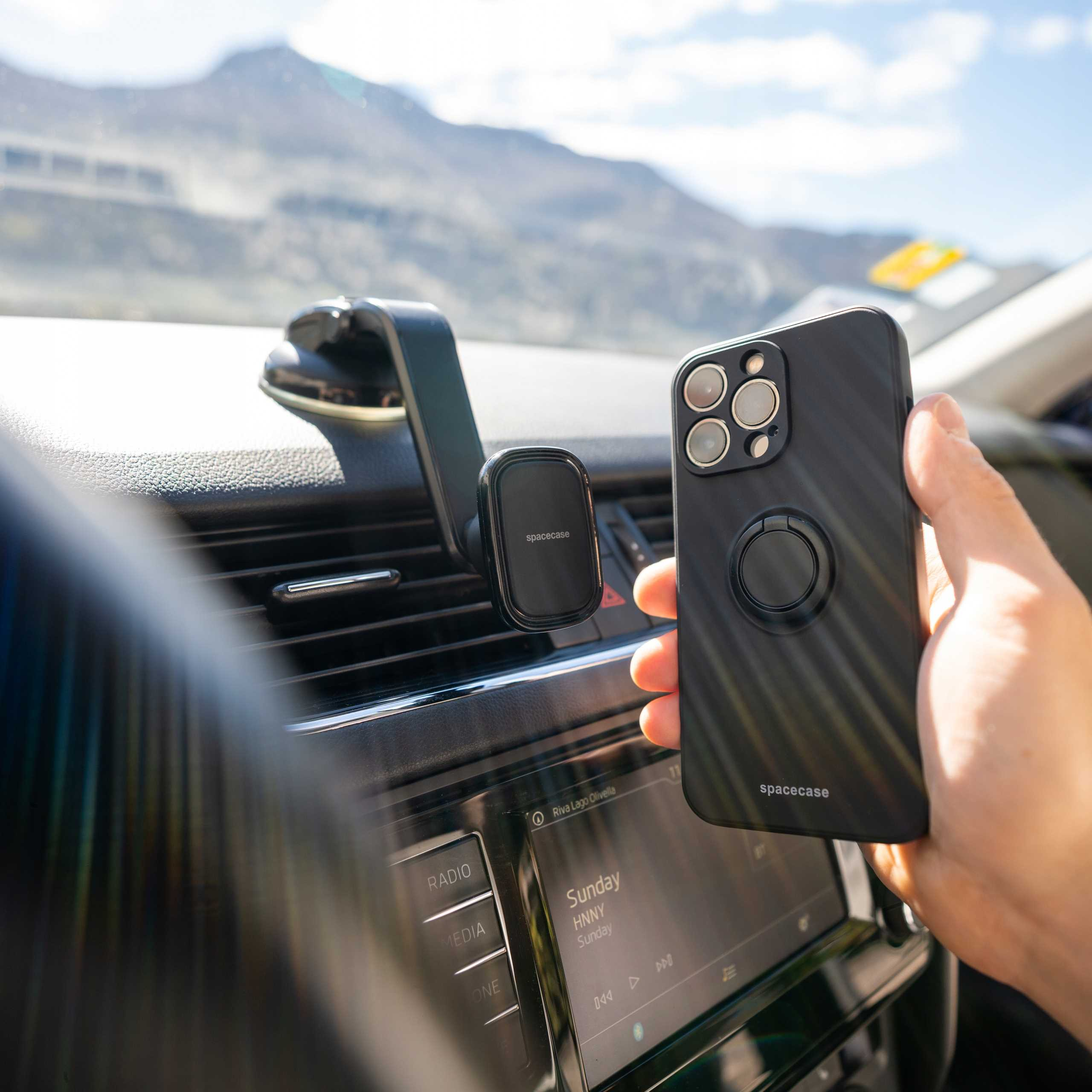 Osoba trzymająca czarny telefon z uchwytem na pierścień w samochodzie. W tle uchwyt i pasmo górskie.