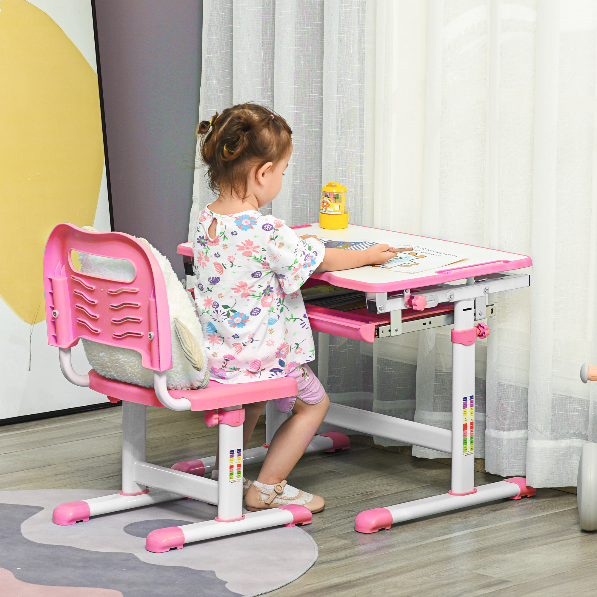 Una bambina è seduta a una scrivania rosa, che guarda un libro. Un contenitore giallo è sulla scrivania. Una tenda bianca sullo sfondo.