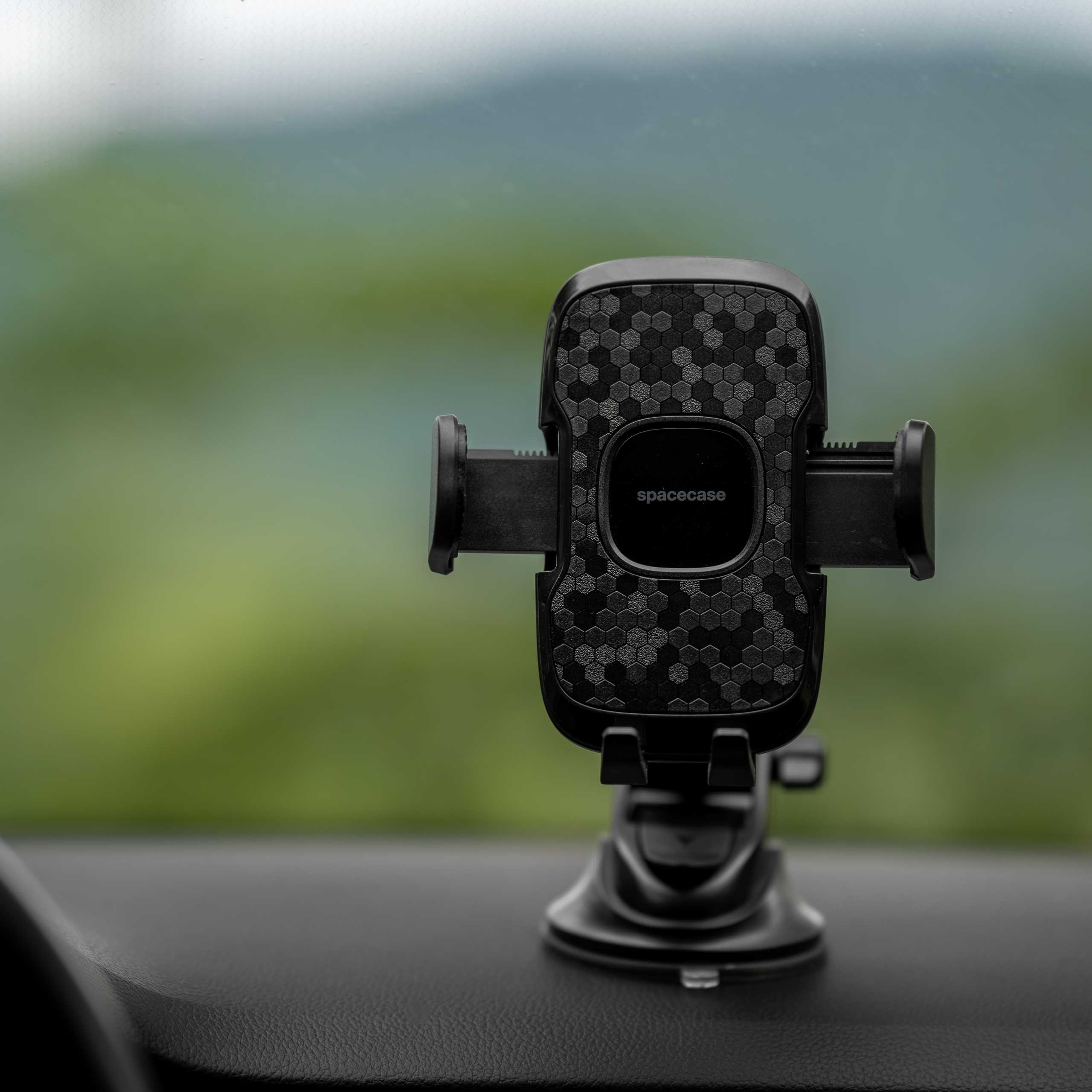 Czarny uchwyt na telefon samochodowy marki 'spacecase' na desce rozdzielczej. Rozmyte zielone tło.