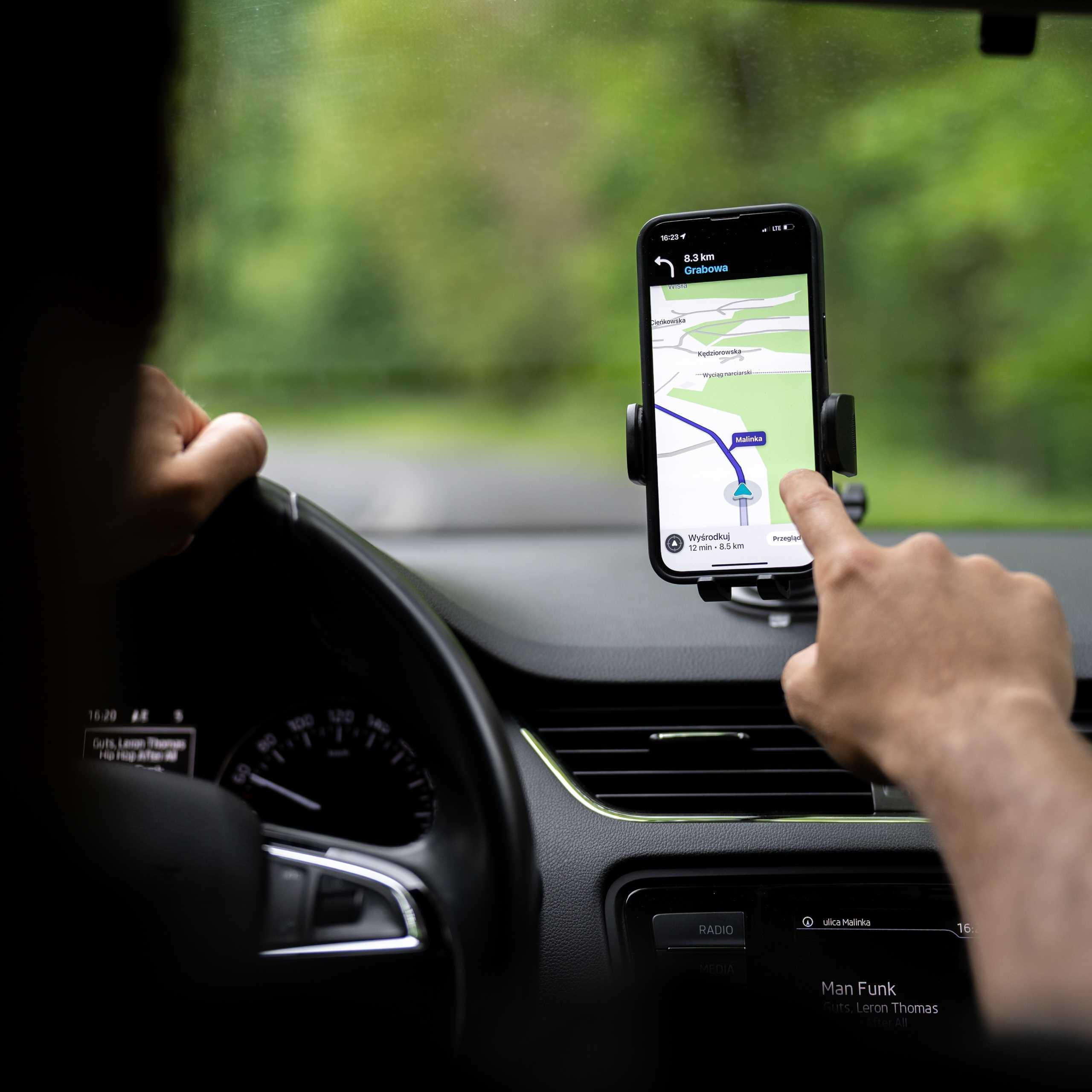 Osoba prowadzi samochód, używając telefonu do nawigacji. Ekran pokazuje mapę. Ręce na kierownicy i telefonie.