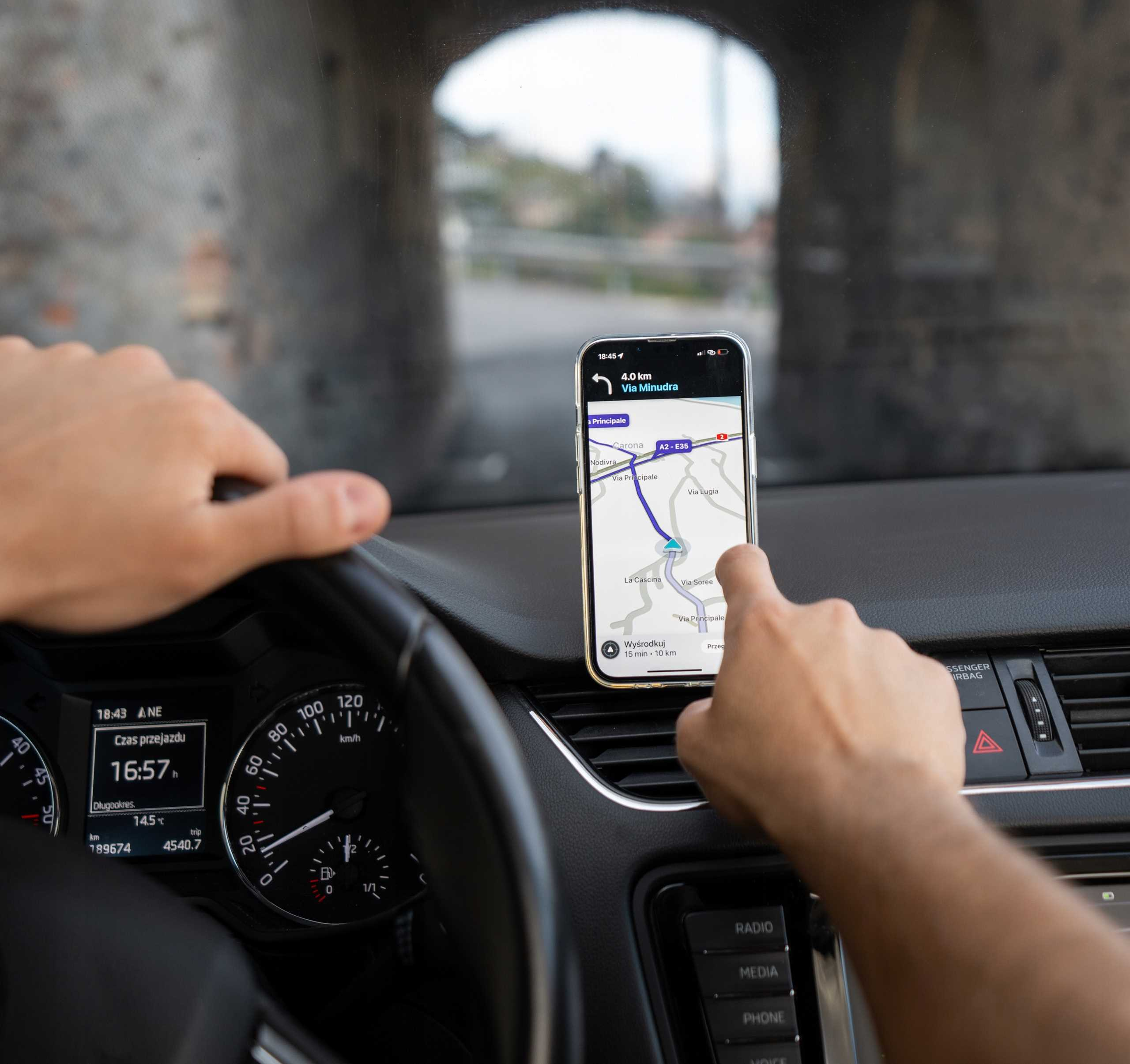 Kierowca używa telefonu do nawigacji. Telefon wyświetla szczegóły trasy. Wnętrze samochodu jest widoczne.