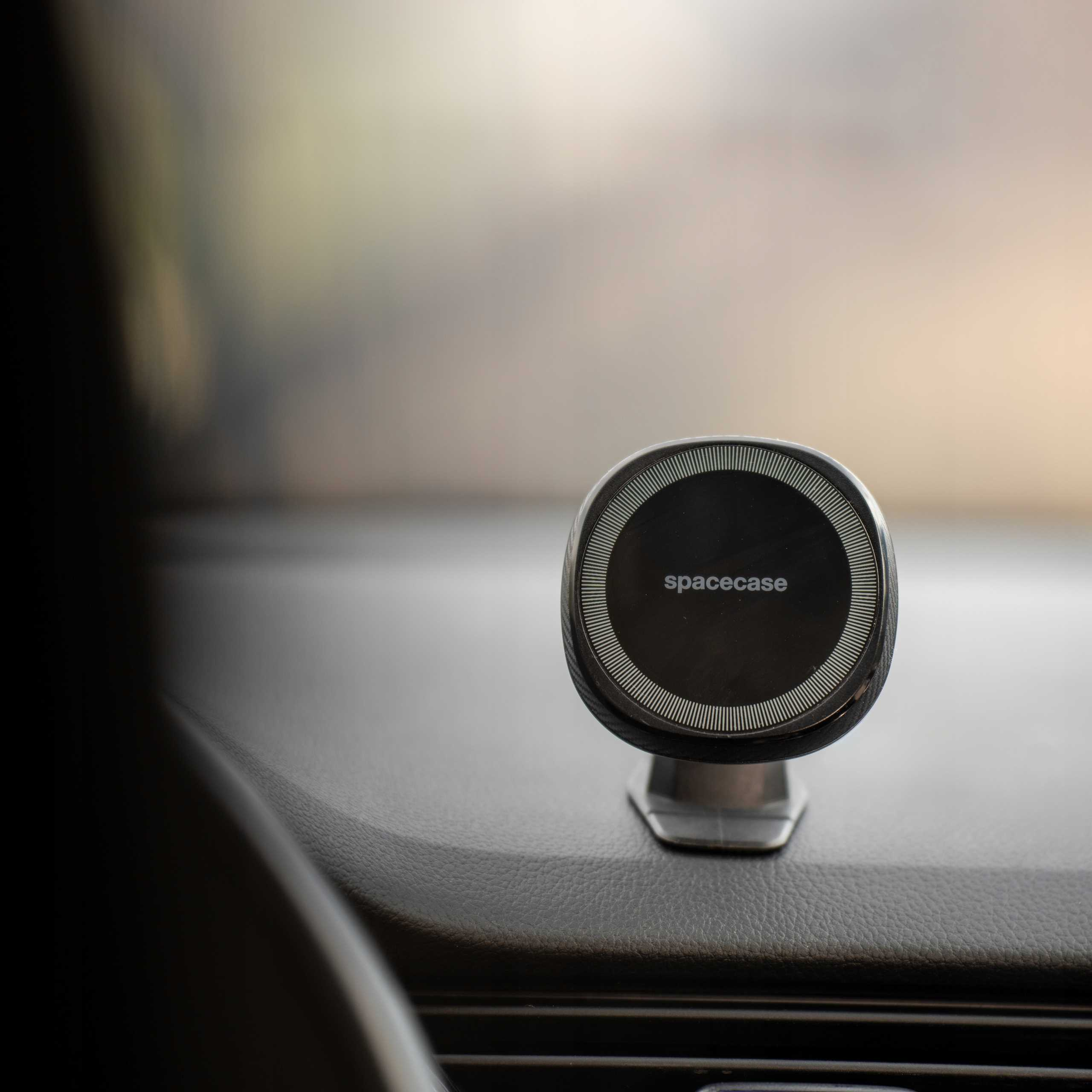 Czarny uchwyt na telefon z napisem 'spacecase' jest zamontowany na desce rozdzielczej samochodu. Tło jest rozmyte.