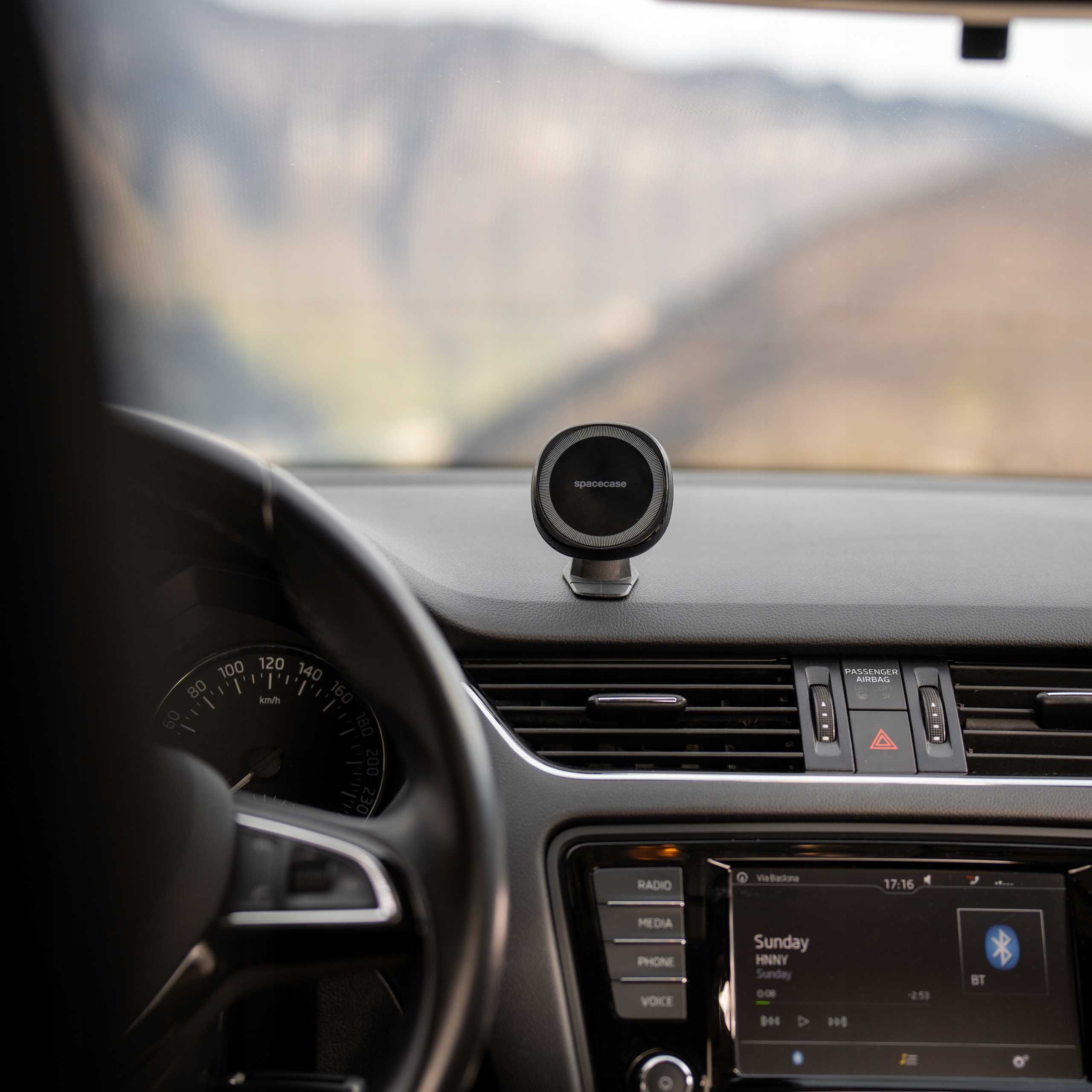 Wnętrze samochodu z uchwytem na telefon na desce rozdzielczej. Widoczny prędkościomierz, radio i kierownica. Góry w tle.