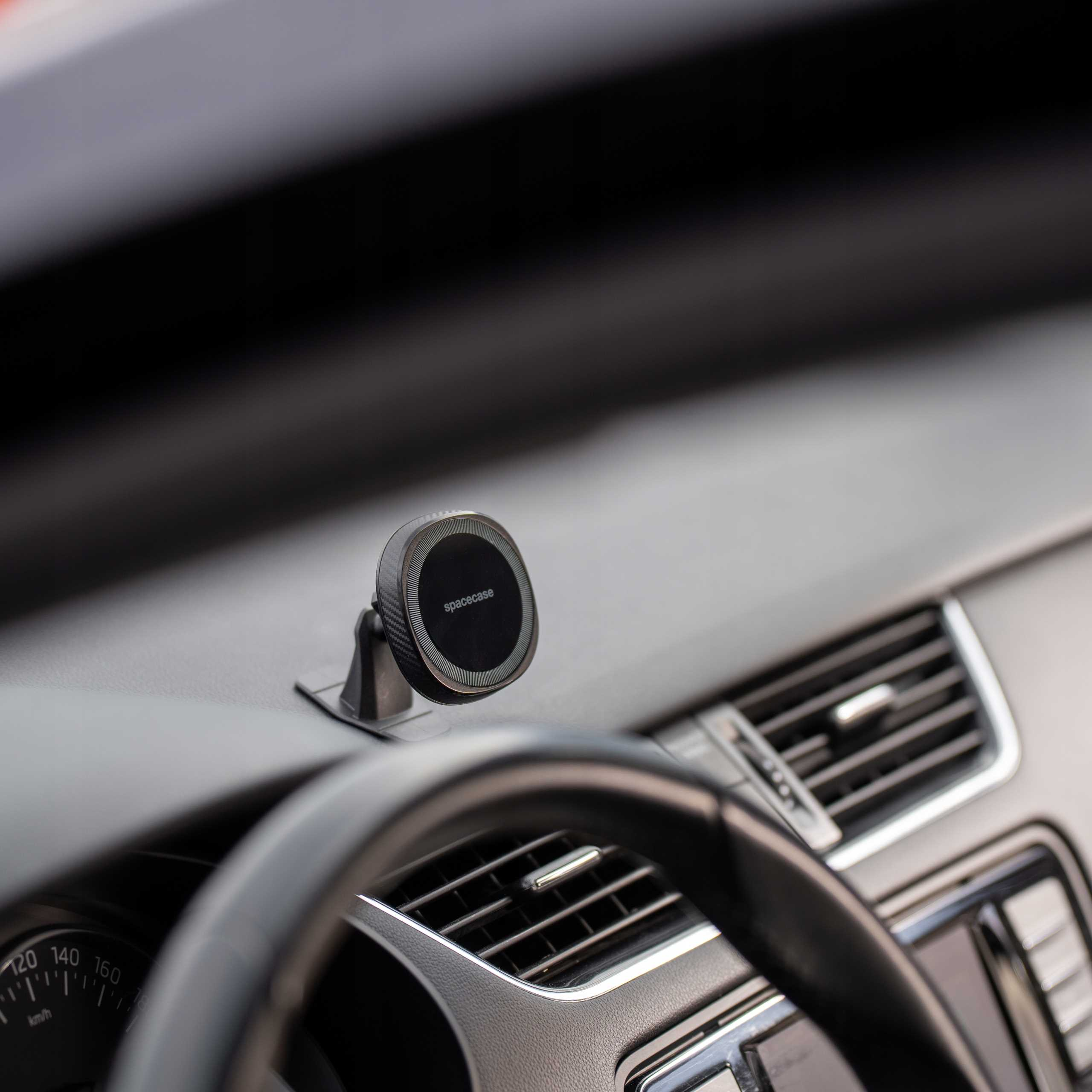 Wnętrze samochodu z uchwytem na telefon. Deska rozdzielcza z otworami wentylacyjnymi i widocznym prędkościomierzem. Kierownica z przodu.