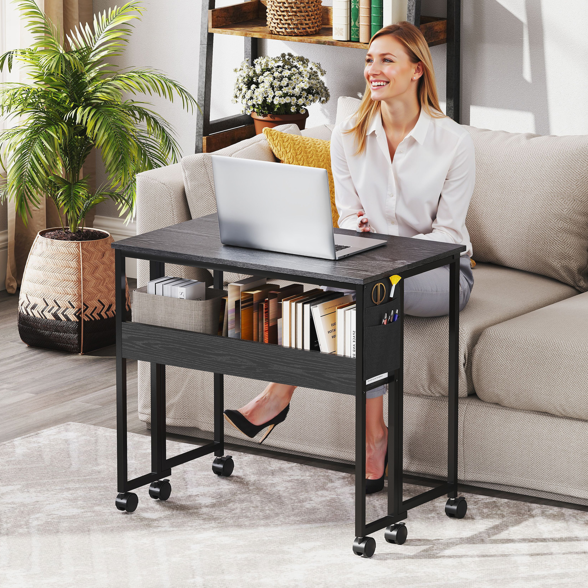 Donna seduta su un divano con laptop su un tavolo con rotelle. Pianta, libri e scaffale sullo sfondo.