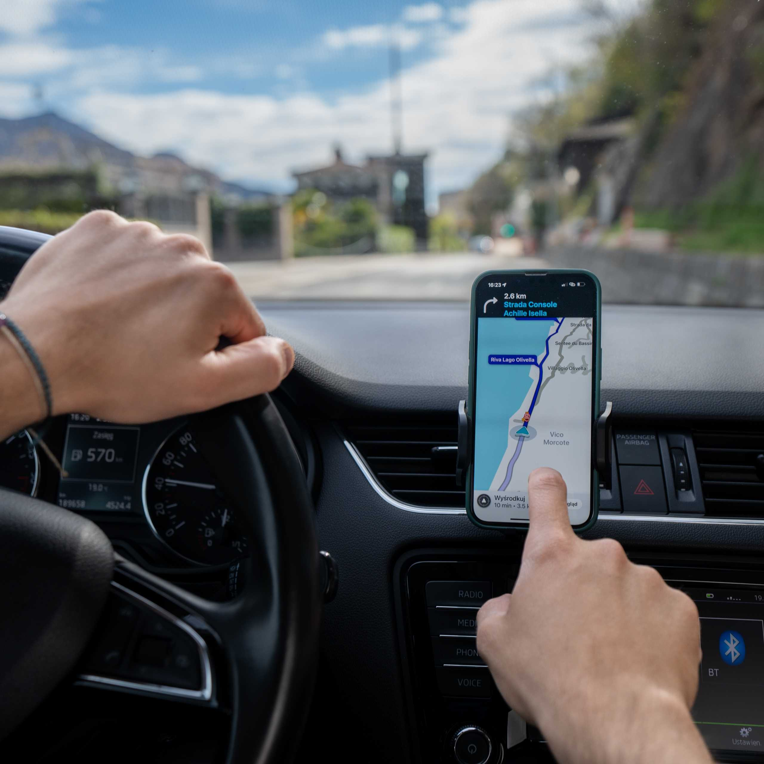 Ręce na kierownicy, telefon wyświetla mapę, dłoń kierowcy dotyka ekranu. Wnętrze samochodu z deską rozdzielczą, Bluetooth i przyciskami radia.