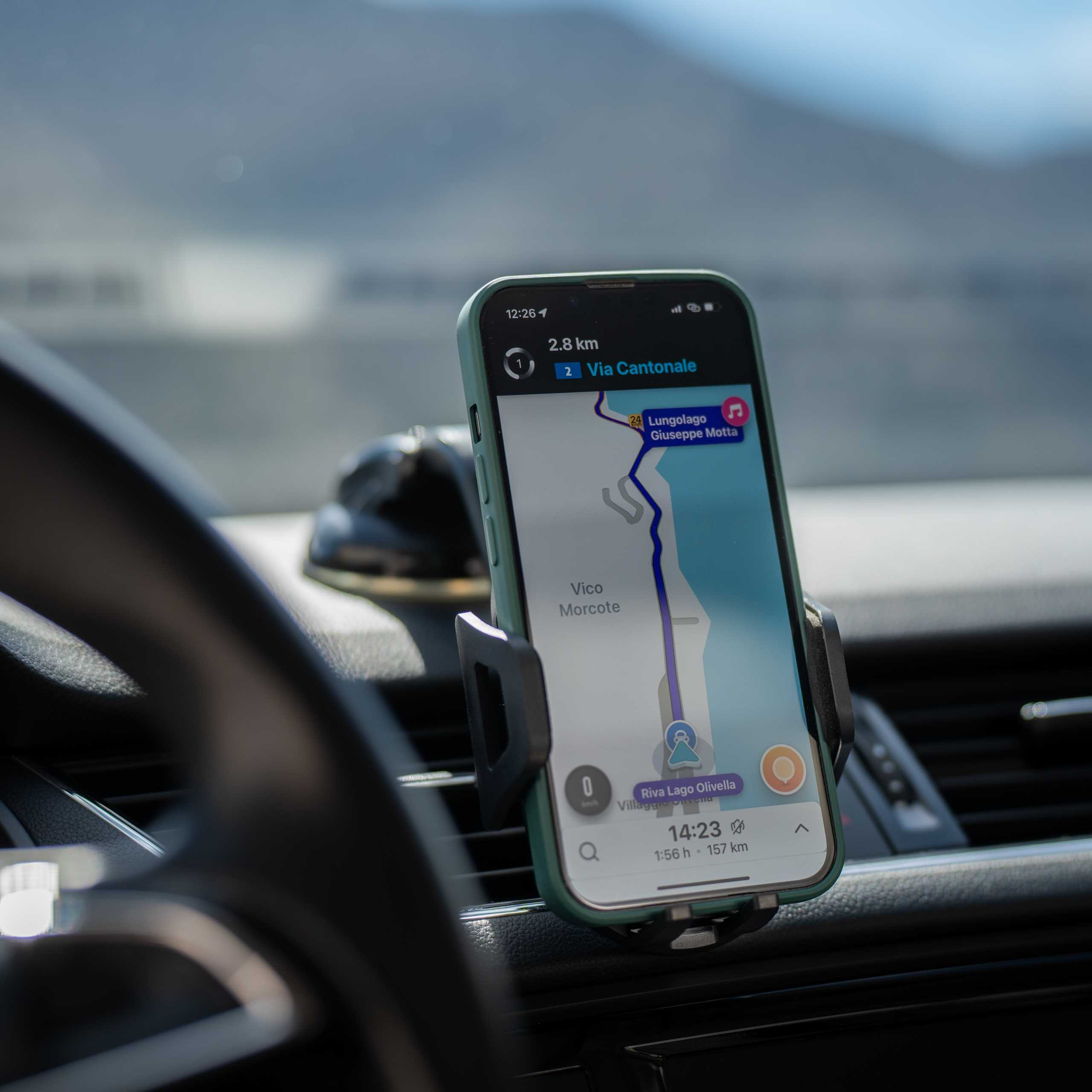Telefon wyświetla mapę nawigacji, umieszczony w uchwycie samochodowym. Widoczna jest deska rozdzielcza i kierownica, a także rozmyte niebo.