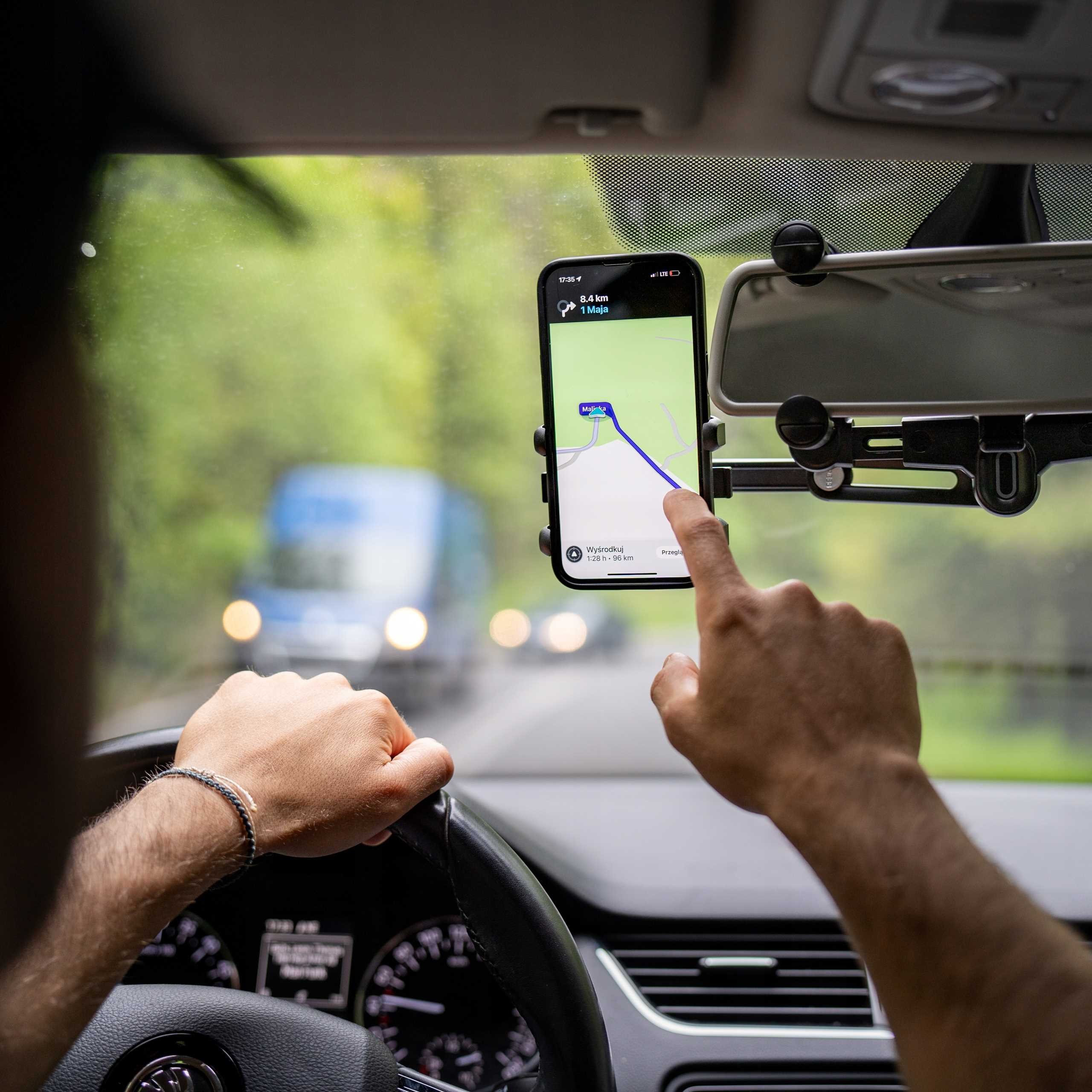 Osoba używająca nawigacji w telefonie w samochodzie. Ręce na kierownicy i dotykające ekranu ze szczegółami trasy.