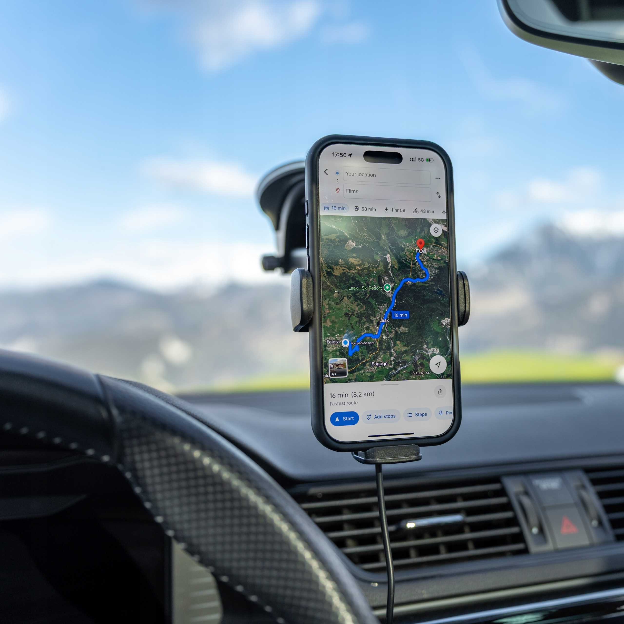Telefon w uchwycie samochodowym pokazuje aplikację nawigacyjną z niebieską trasą na mapie. Widoczne góry i niebo.