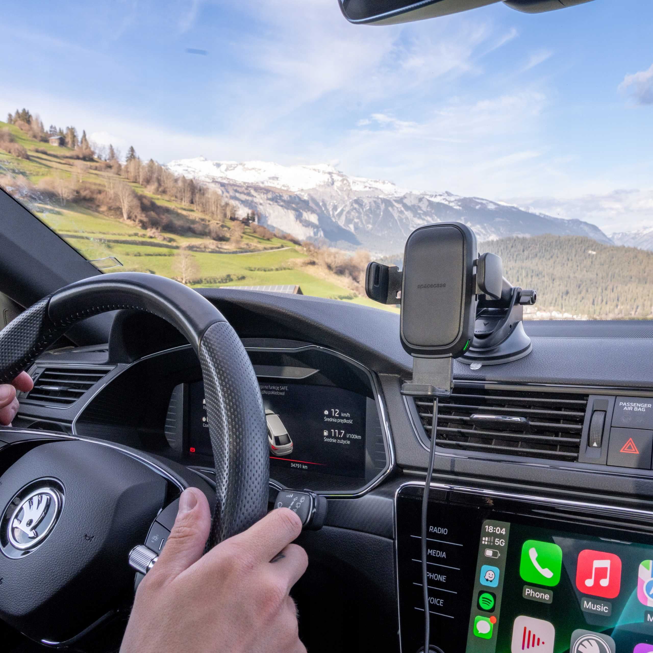 Widok wnętrza samochodu. Kierownica, deska rozdzielcza z uchwytem na telefon i widok gór oraz zielonych wzgórz.
