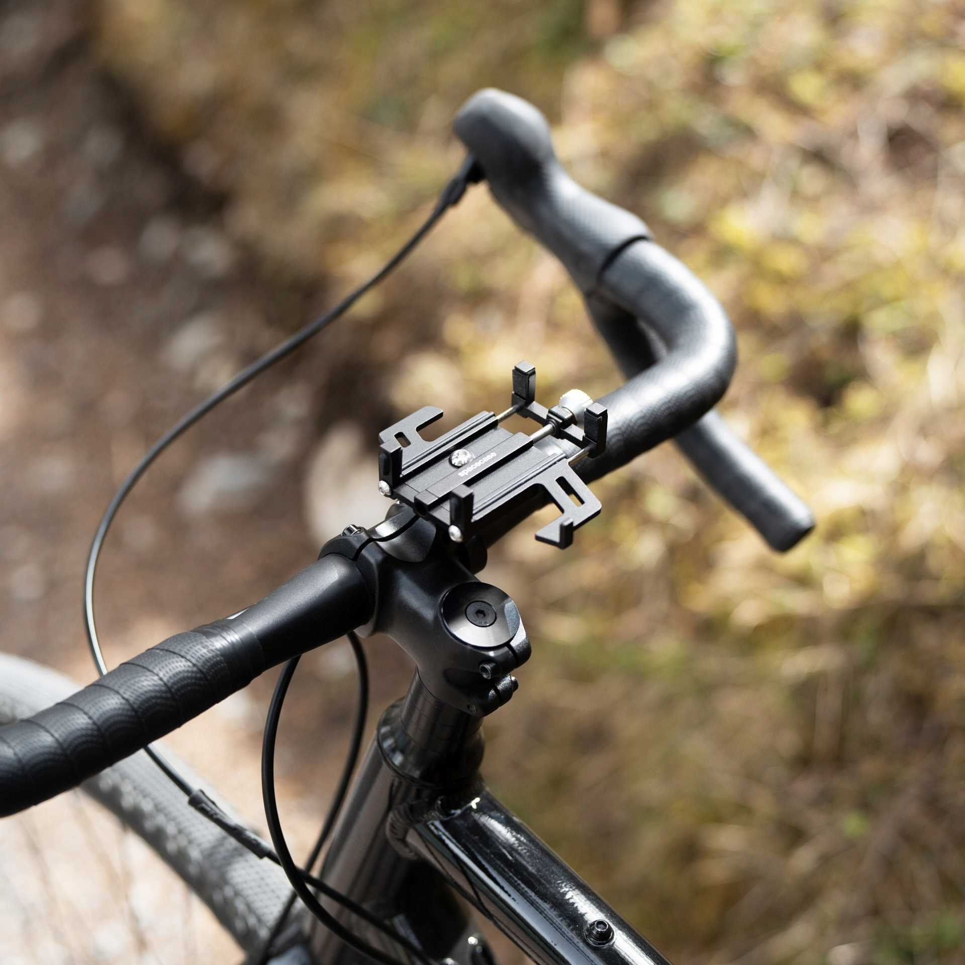 Czarna kierownica roweru z uchwytem na telefon. Tło jest rozmyte, w naturalnym, ziemistym odcieniu.