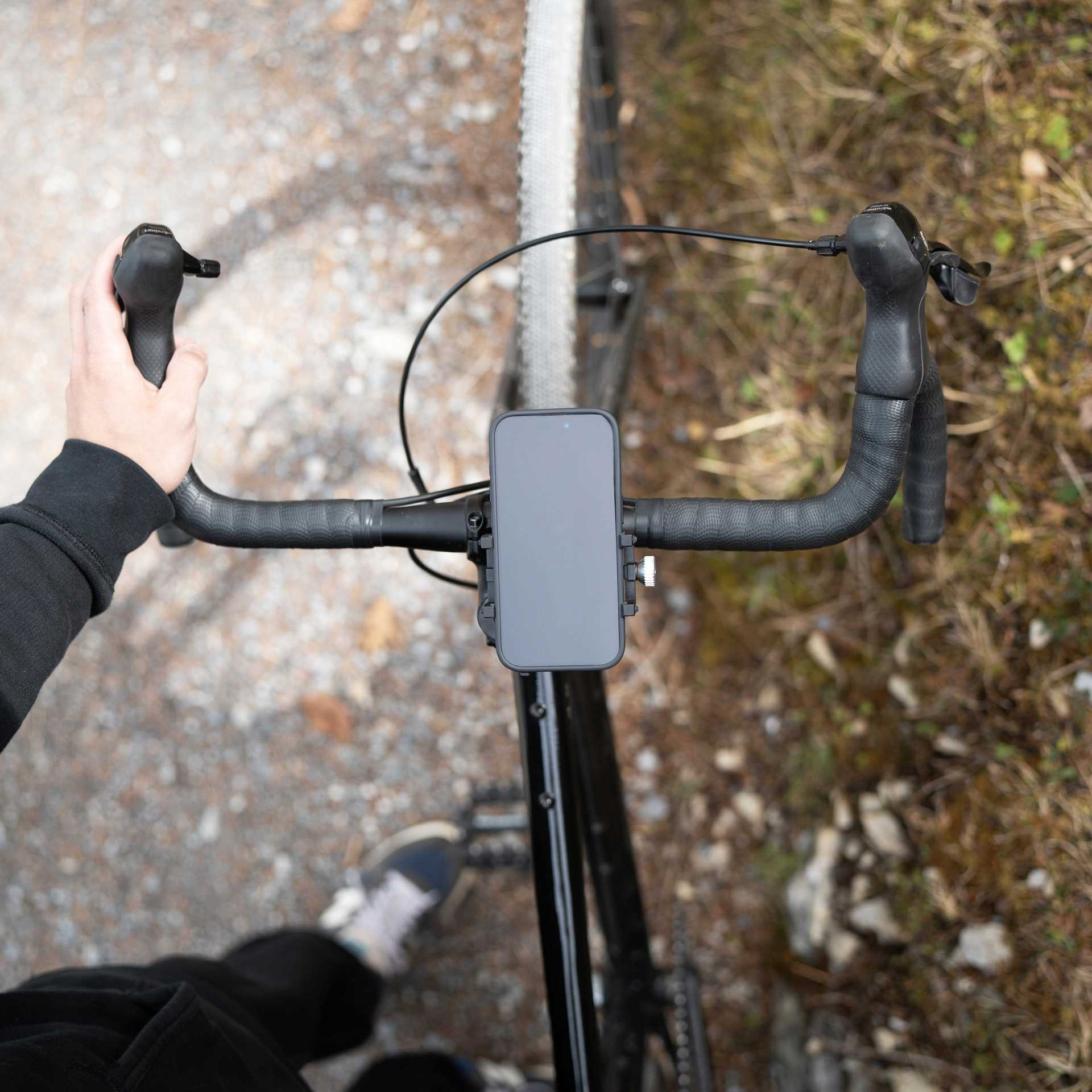 Widok z góry na kierownicę roweru z uchwytem na telefon. Lewa ręka jest widoczna. Widoczny jest grunt.