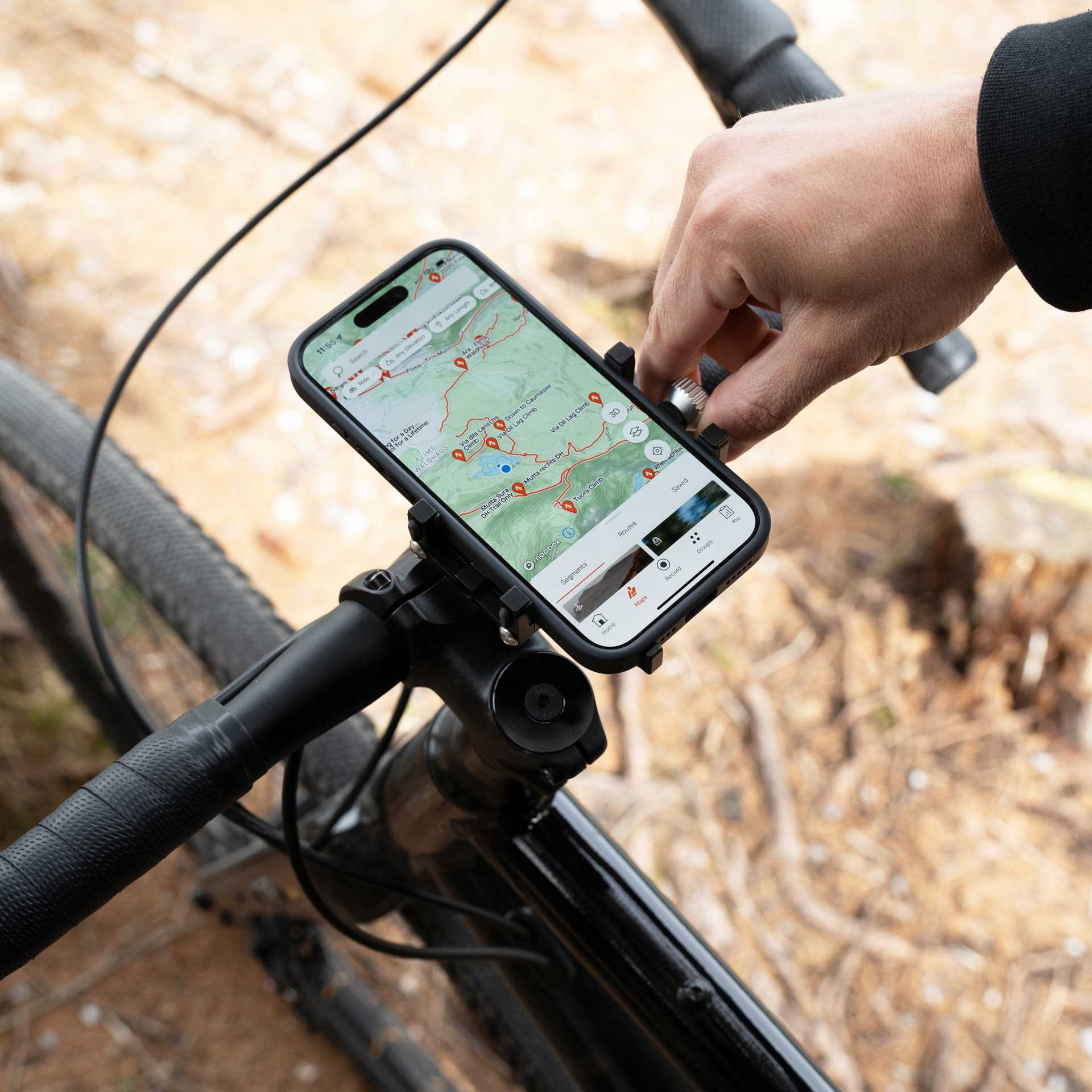 Ręka reguluje telefon na kierownicy roweru. Telefon wyświetla mapę. Rower jest czarny. Tło to rozmyty las.