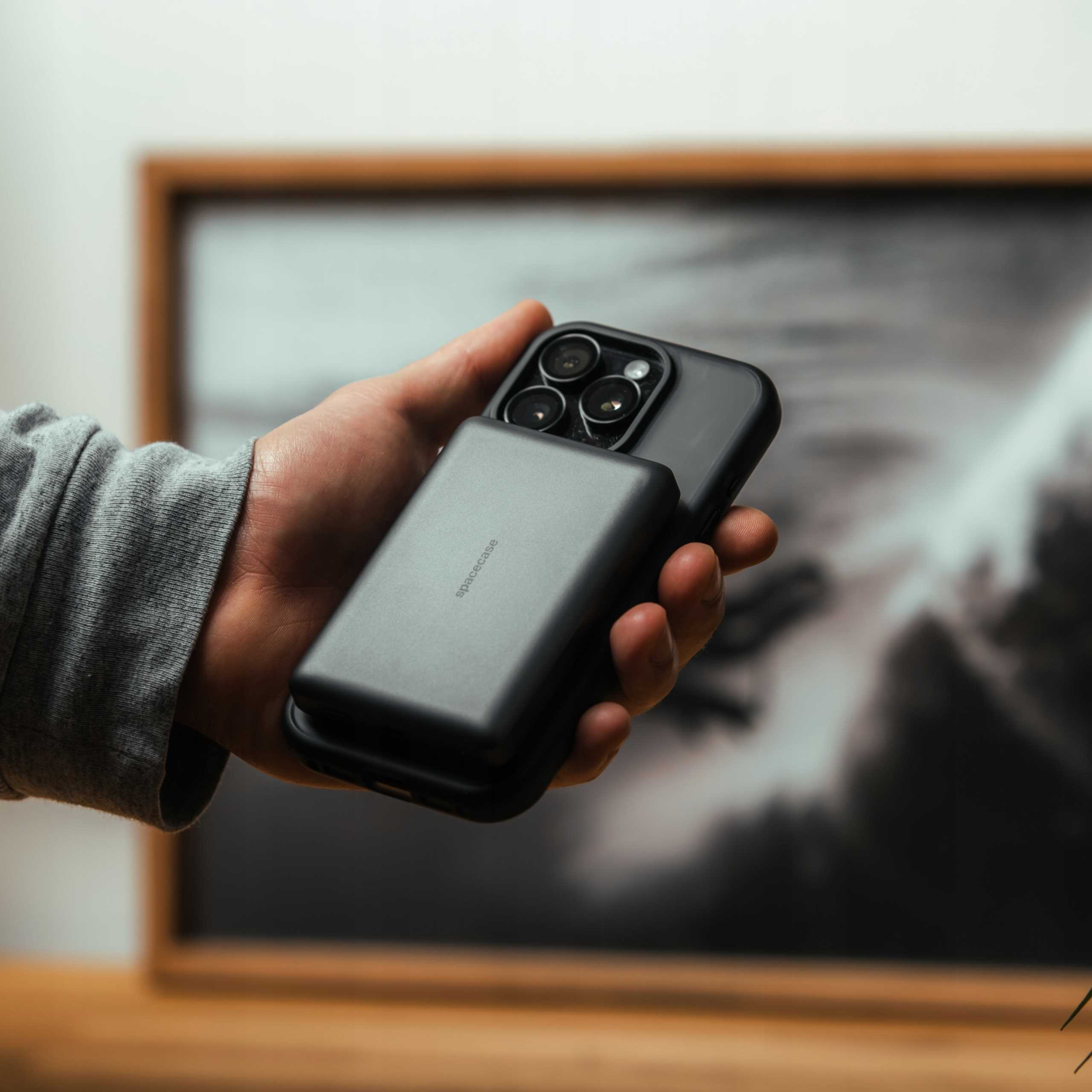 Dłoń trzyma czarny telefon z nakładką na aparat. W tle wisi oprawiony obraz.