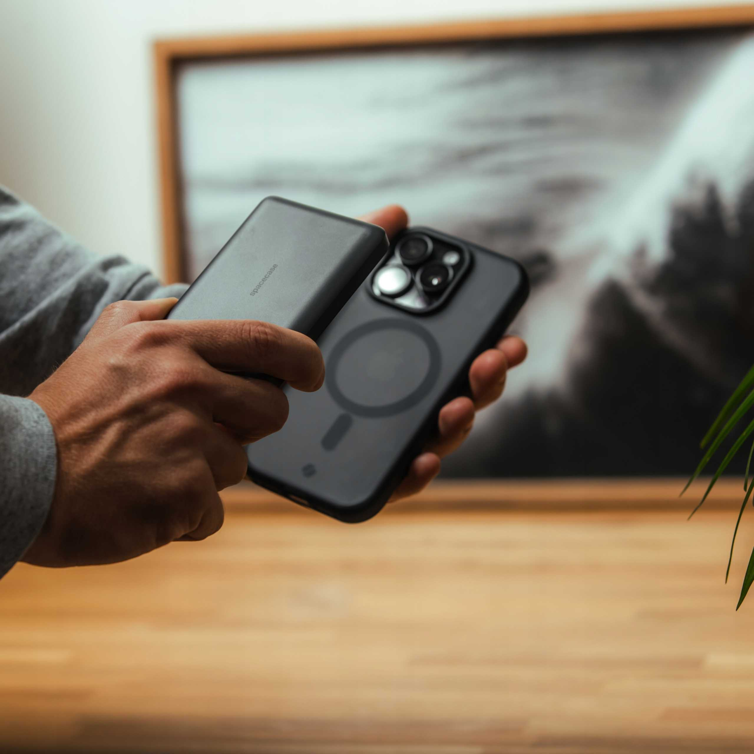 Ręce trzymają telefon i power bank. Telefon jest czarny i ma 3 aparaty. Power bank jest szary.