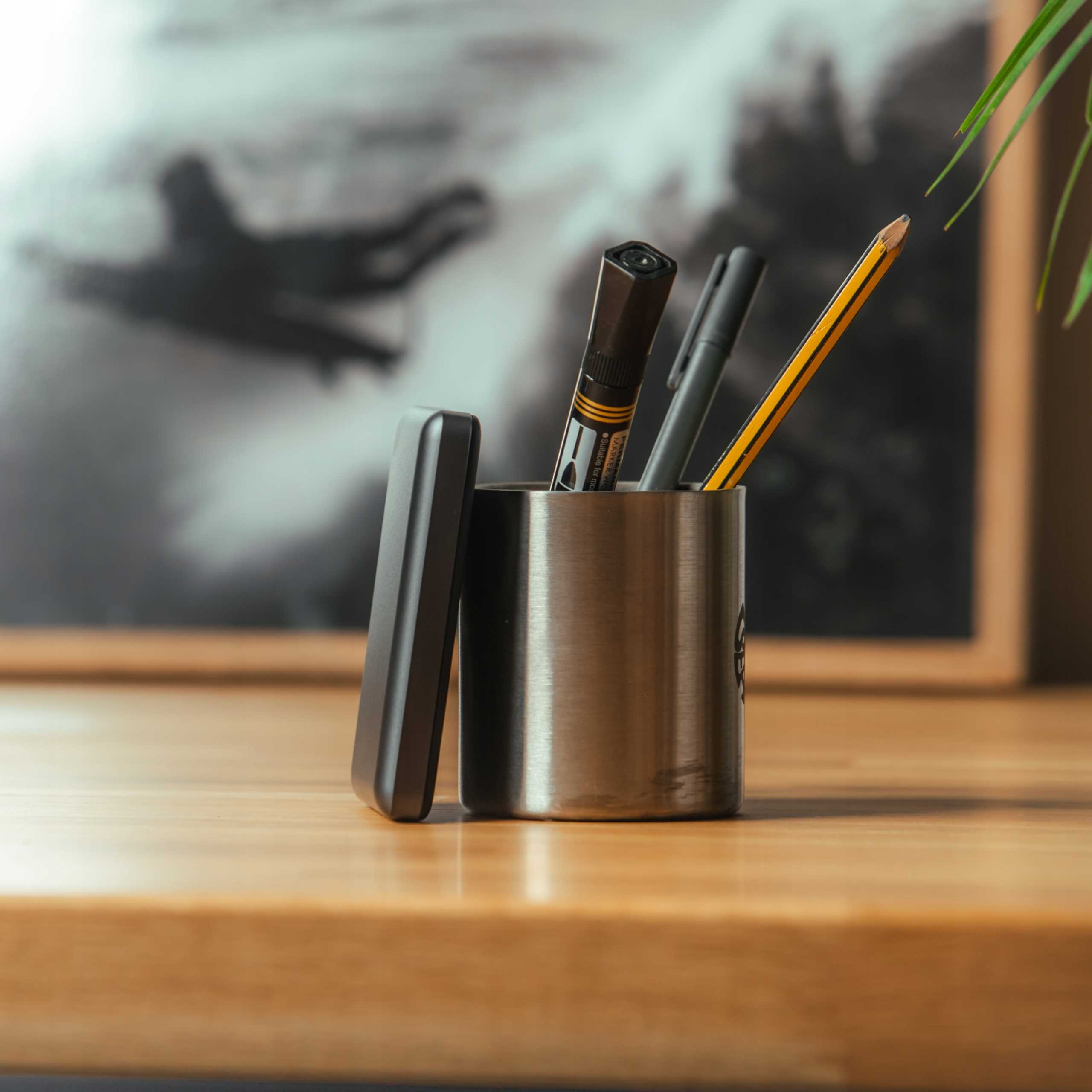 Metalowy pojemnik na długopisy z długopisami i ołówkami stoi na drewnianej powierzchni. Rozmyte zdjęcie jest w tle.