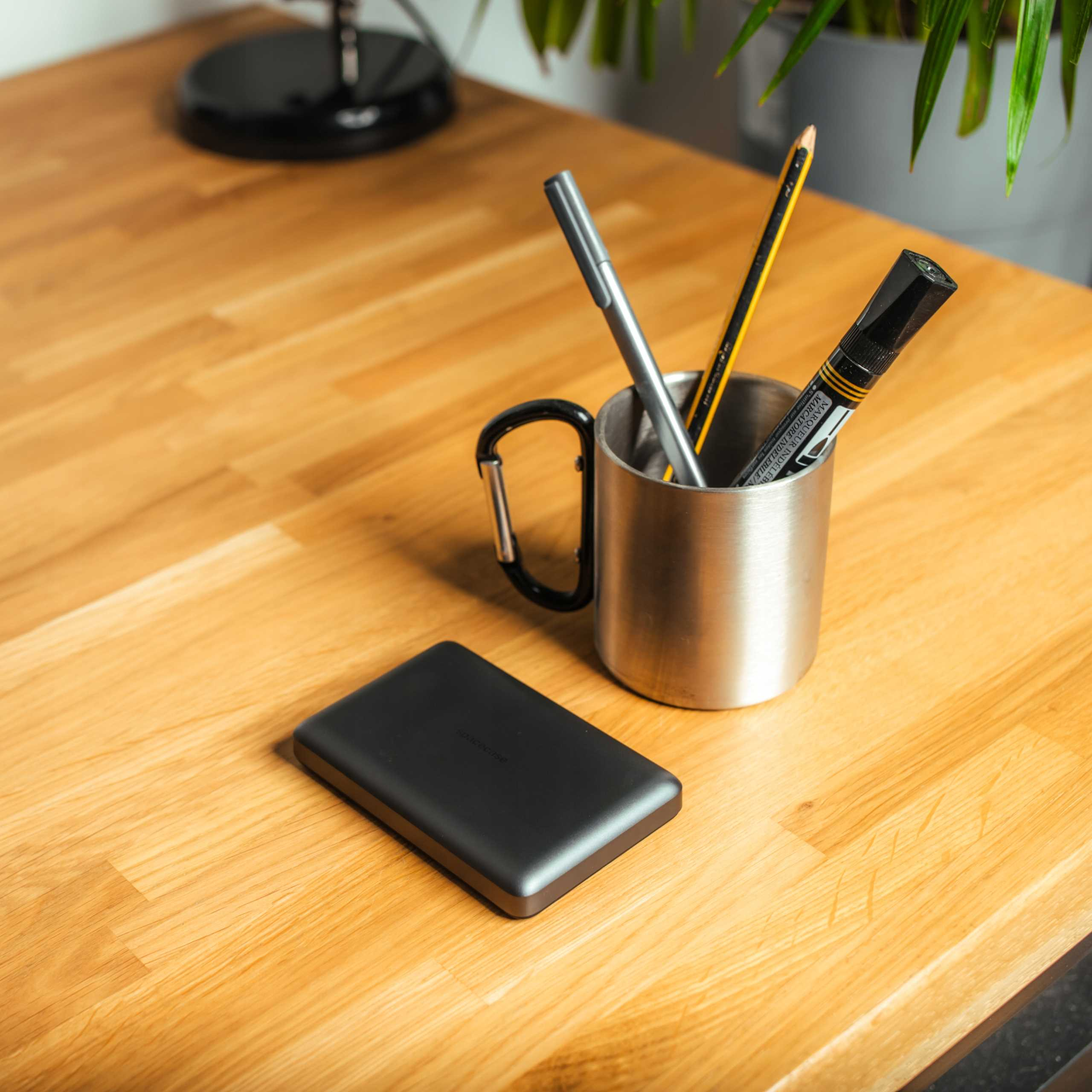 Drewniane biurko z długopisem, ołówkiem i markerem w metalowym kubku. Obok leży czarny zewnętrzny dysk twardy.