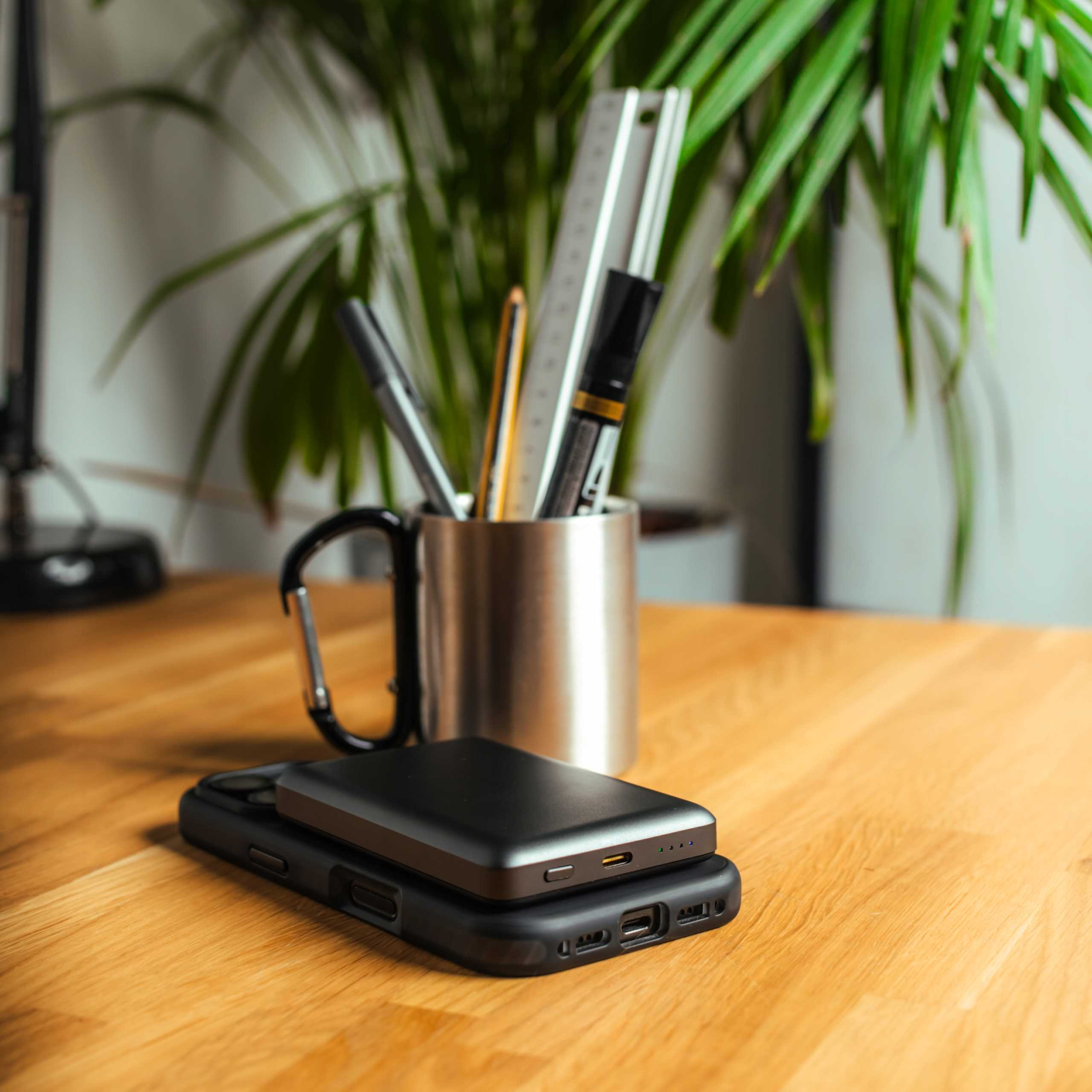 Drewniane biurko z etui na telefon i power bankiem. Metalowy kubek z długopisami i linijką z tyłu.