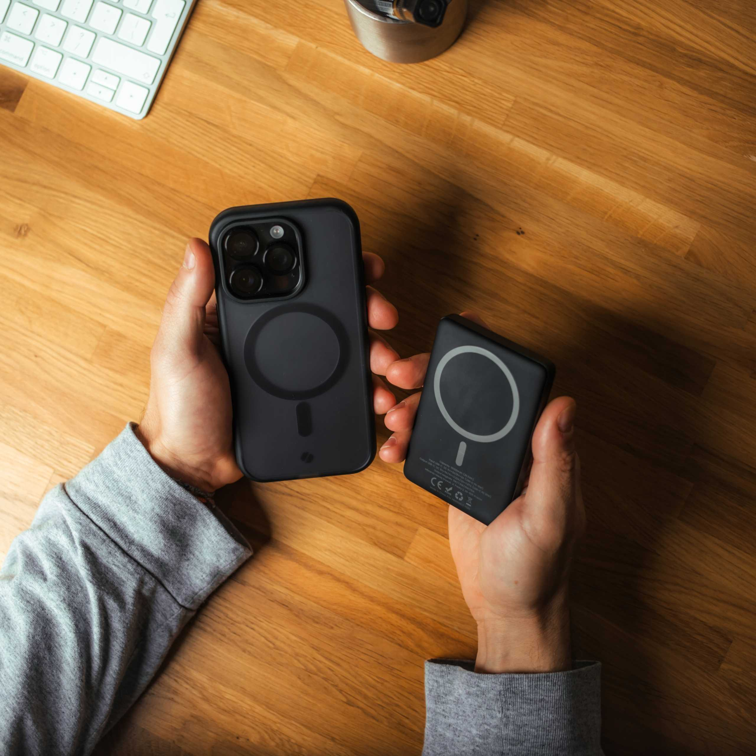 Dłonie trzymające czarnego iPhone'a i czarną przenośną ładowarkę na drewnianym biurku. W tle klawiatura i pojemnik na długopisy.