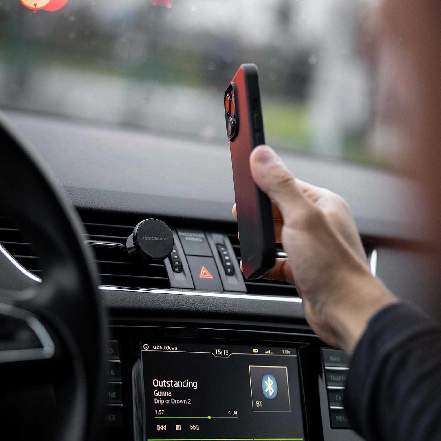 Osoba trzymająca smartfon w samochodzie, blisko deski rozdzielczej. Telefon ma czerwono-czarne etui. Radio samochodowe jest widoczne.