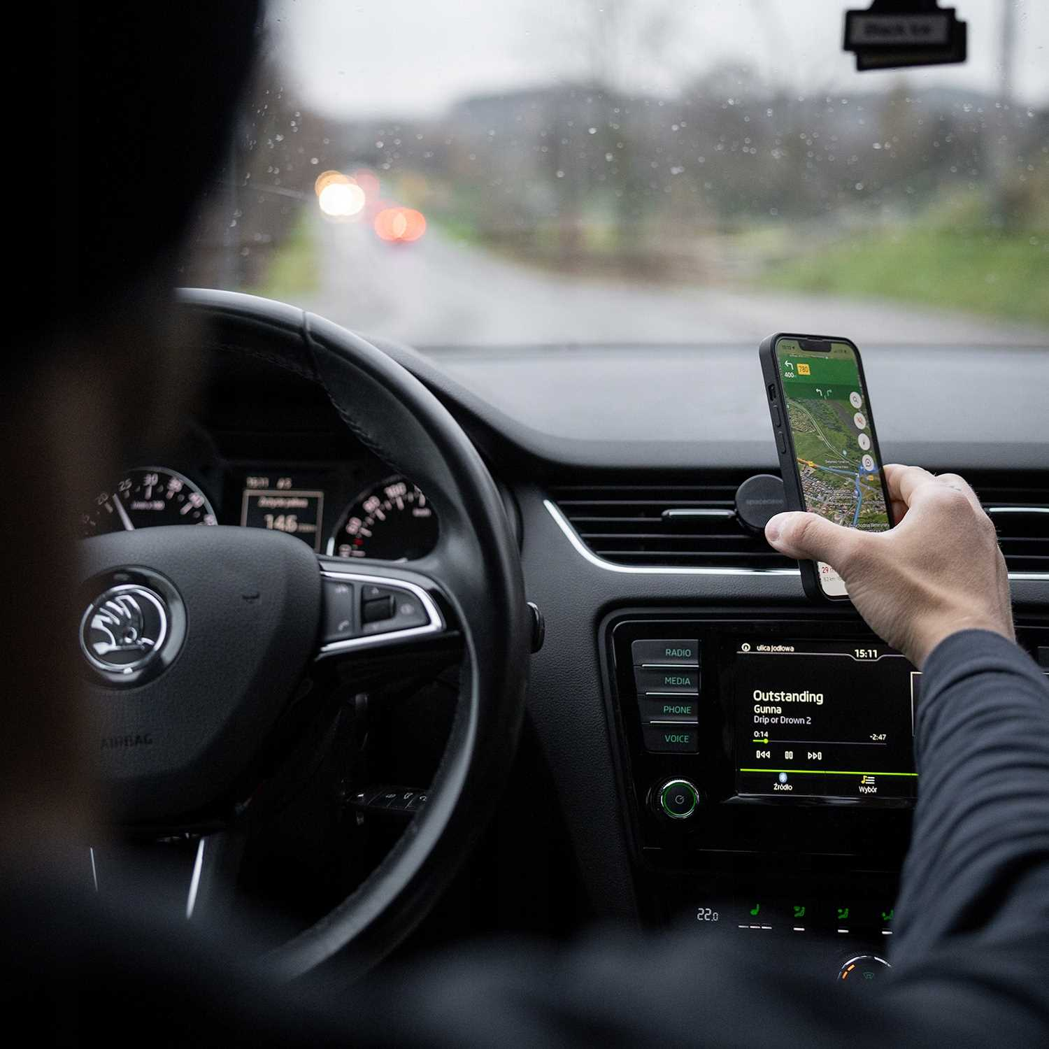 Osoba trzymająca telefon z aplikacją map w samochodzie. Widoczna deska rozdzielcza i kierownica. Droga jest lekko rozmyta.