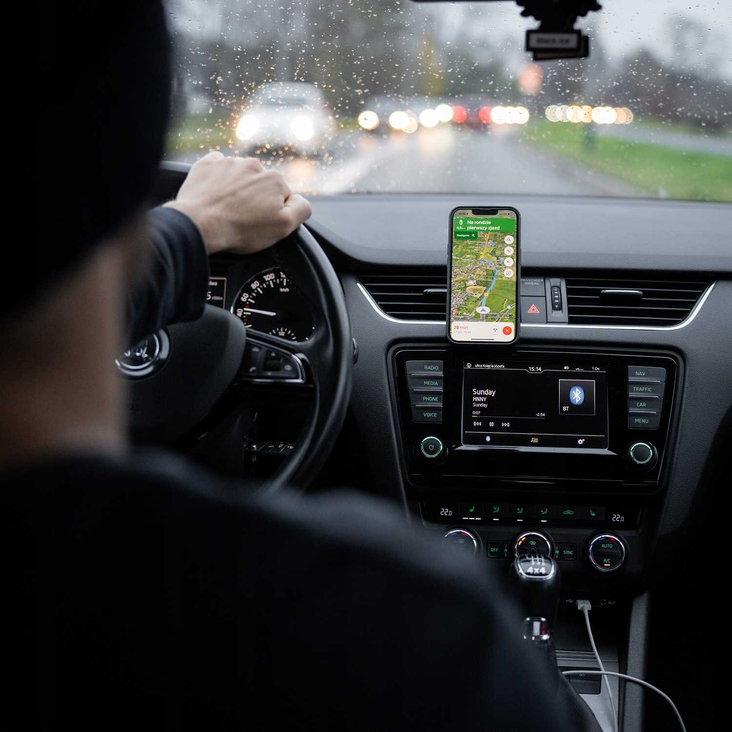 Osoba prowadzi samochód, trzymając kierownicę. Telefon z aplikacją mapy jest zamontowany na desce rozdzielczej.