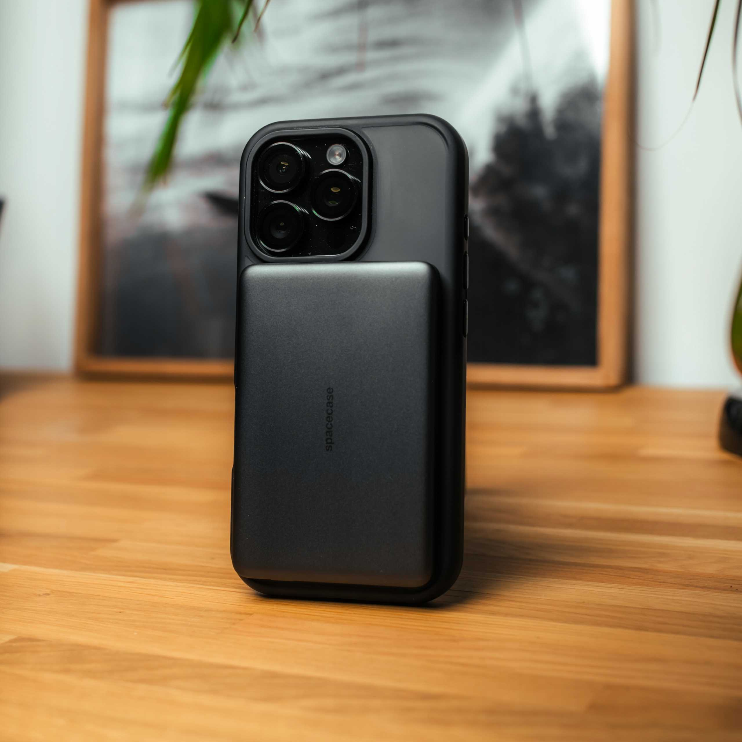 Czarny iPhone z czarnym power bankiem, leżący na drewnianej powierzchni. W tle obraz w ramie i roślina.