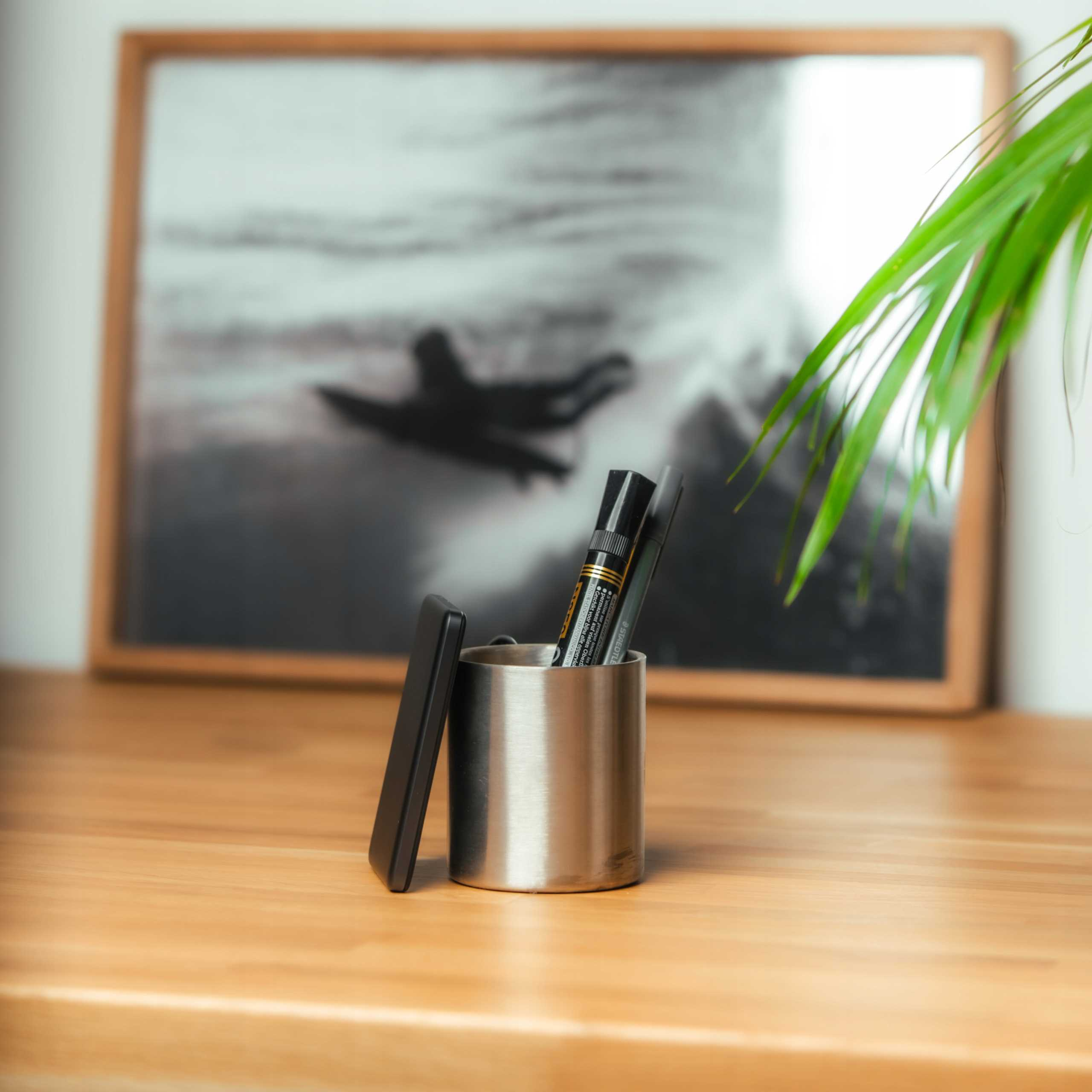 Metalowy piórnik z długopisami i czarną pokrywką stoi na drewnianym stole, na tle oprawionego obrazu i zielonej rośliny.