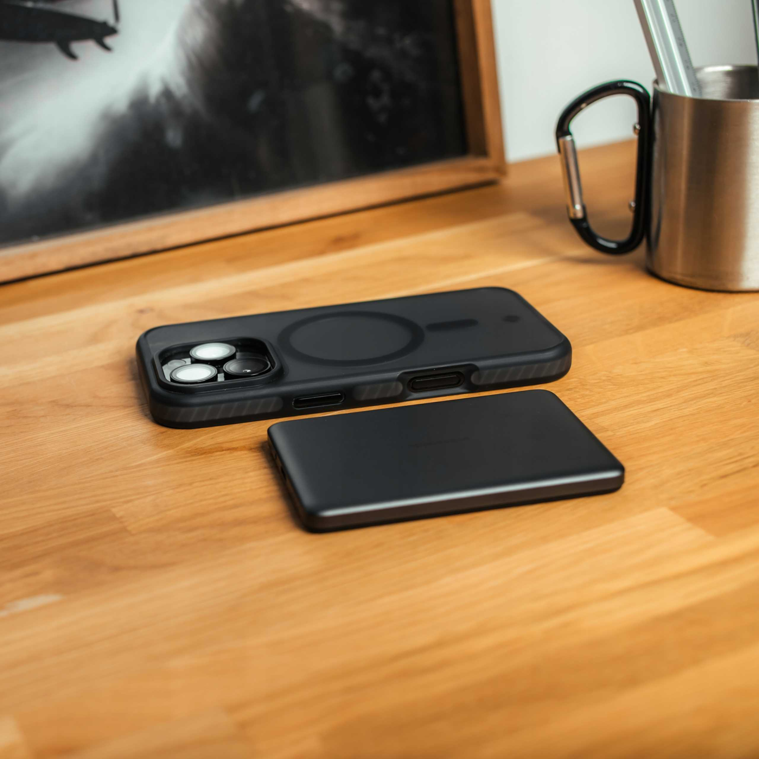 Czarna obudowa telefonu i ładowarka leżą na drewnianym stole, obok oprawionego zdjęcia i metalowego kubka.