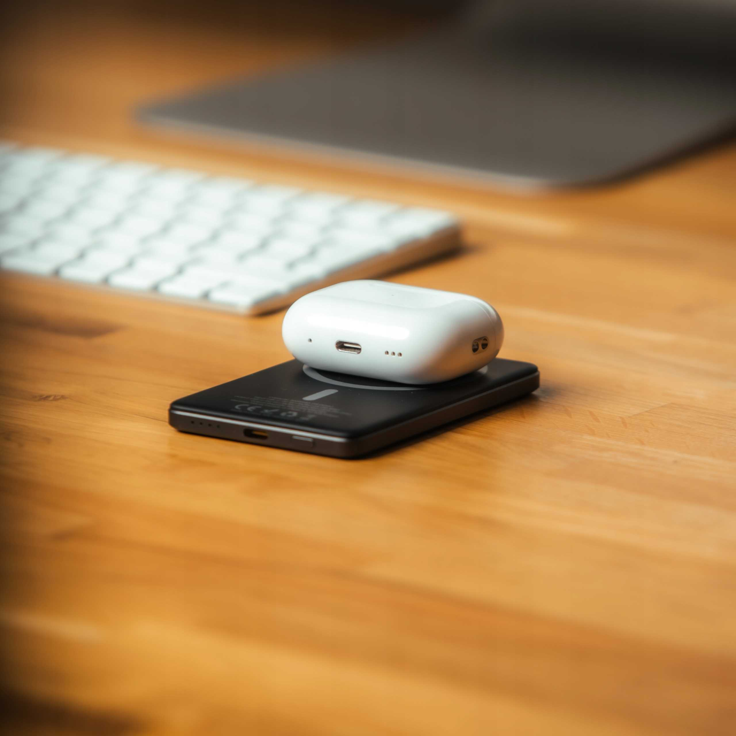 Białe słuchawki ładujące się na czarnej ładowarce bezprzewodowej, na drewnianym biurku. Biała klawiatura w tle.