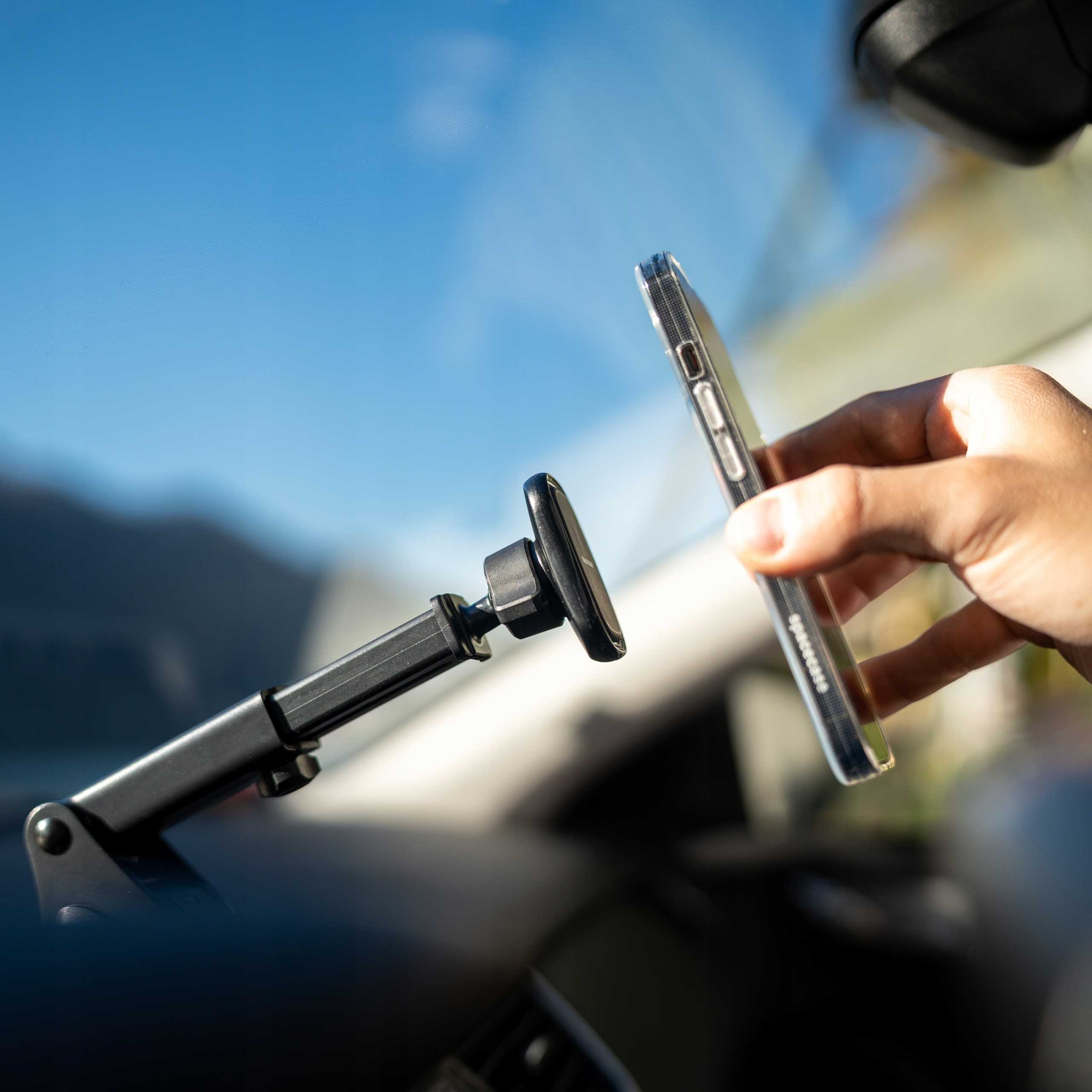 Ręka trzymająca telefon w samochodzie, w pobliżu uchwytu na telefon, z niebieskim niebem widocznym przez przednią szybę.