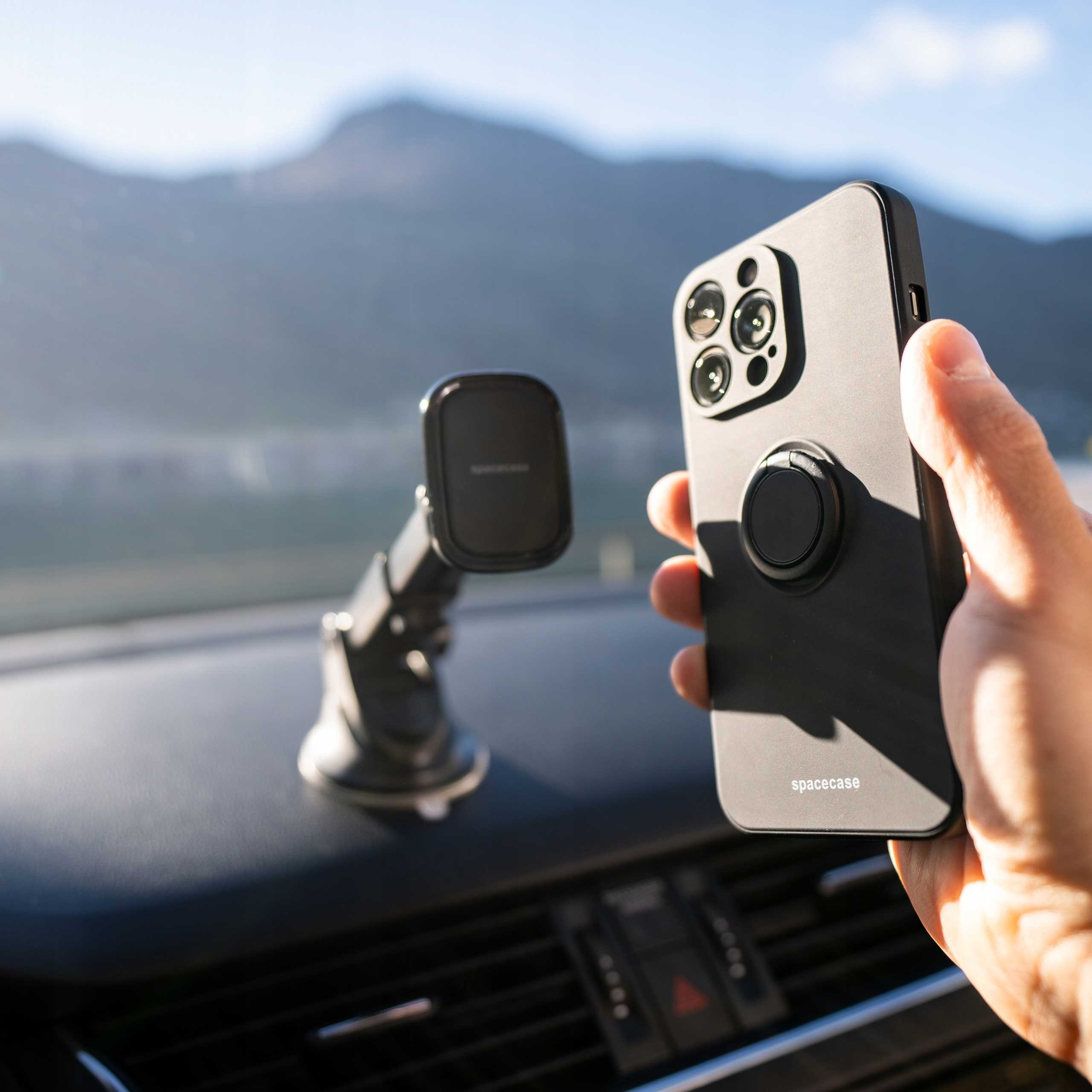 Osoba trzymająca czarny telefon z uchwytem na pierścień w samochodzie. Uchwyt na telefon jest na desce rozdzielczej. Góry w tle.