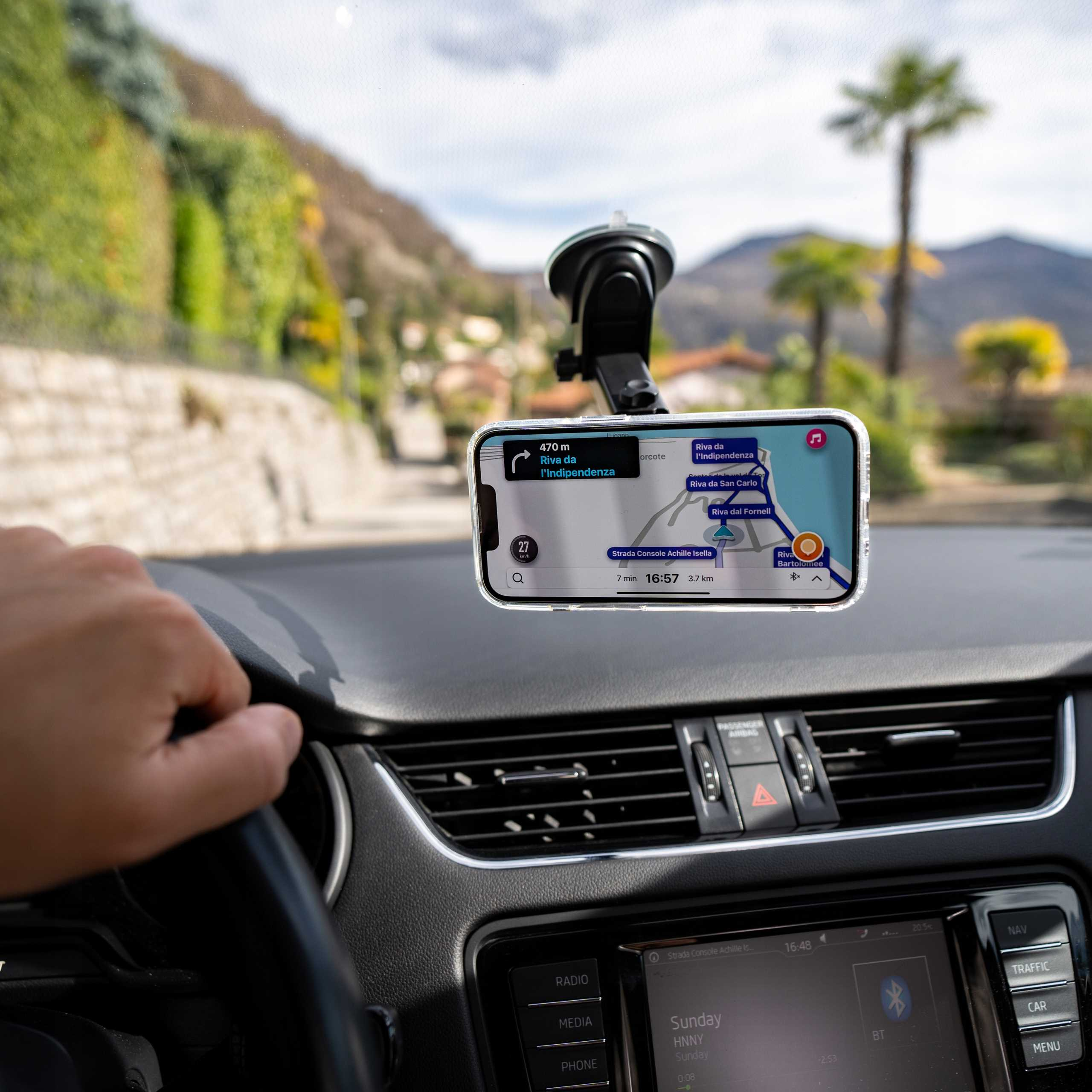 Ręka na kierownicy, telefon z mapą. Zielone wzgórza i palma widoczne przez przednią szybę.