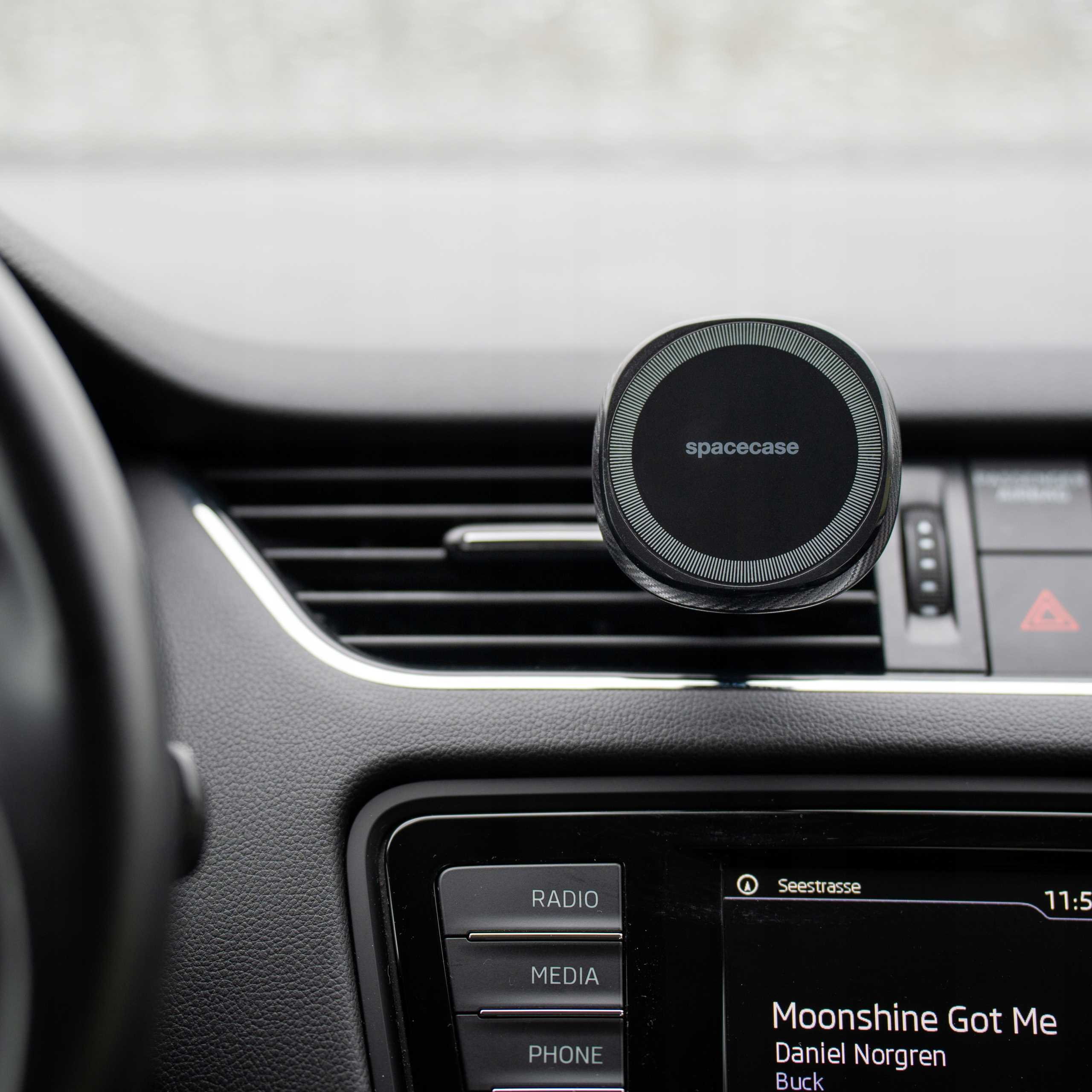 Wnętrze samochodu pokazuje uchwyt na telefon i wyświetlacz radia. Uchwyt ma napis 'spacecase'. Wyświetlacz pokazuje piosenkę i stację.