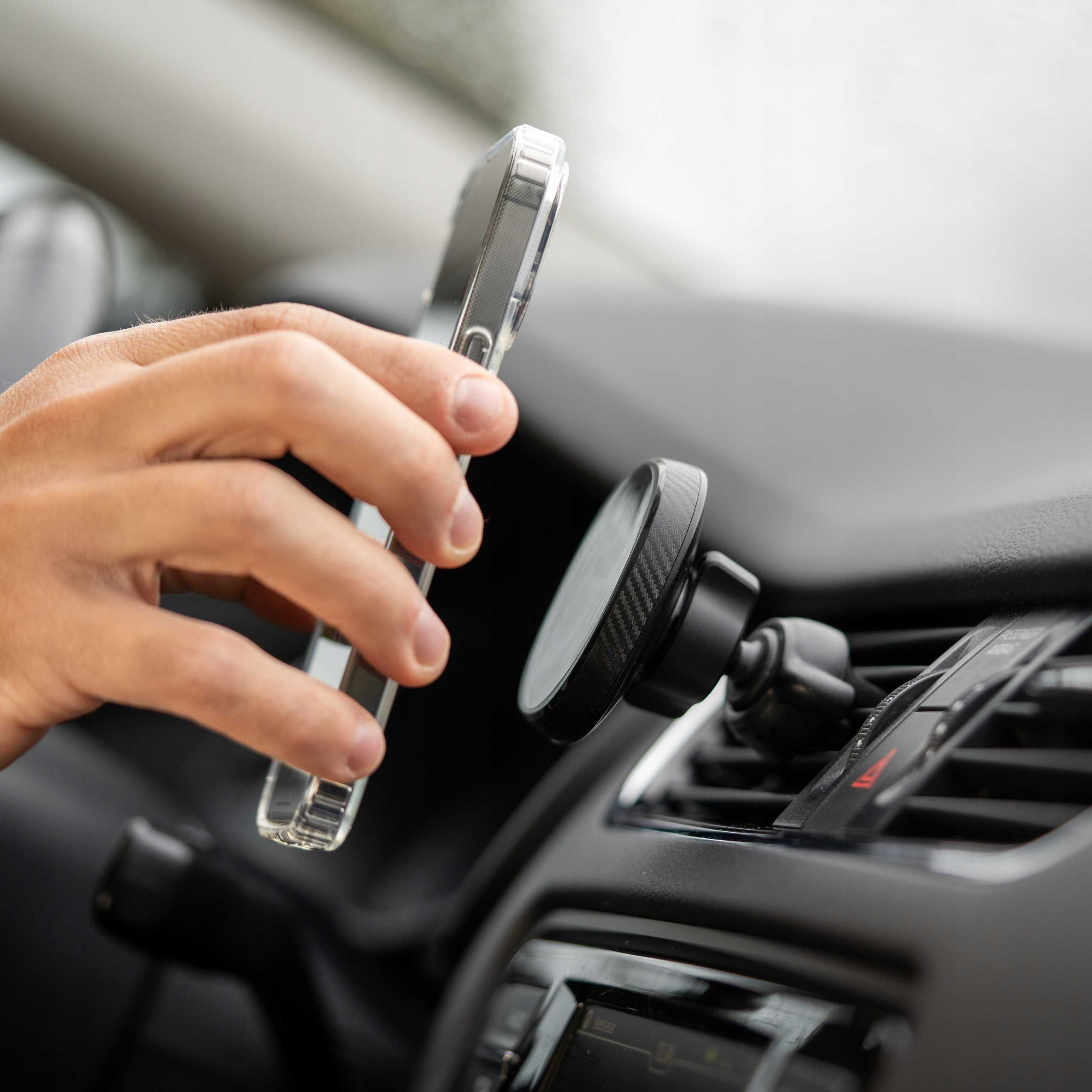 Ręka trzymająca telefon w przezroczystym etui nad uchwytem samochodowym, zamontowanym na wentylacji deski rozdzielczej samochodu.