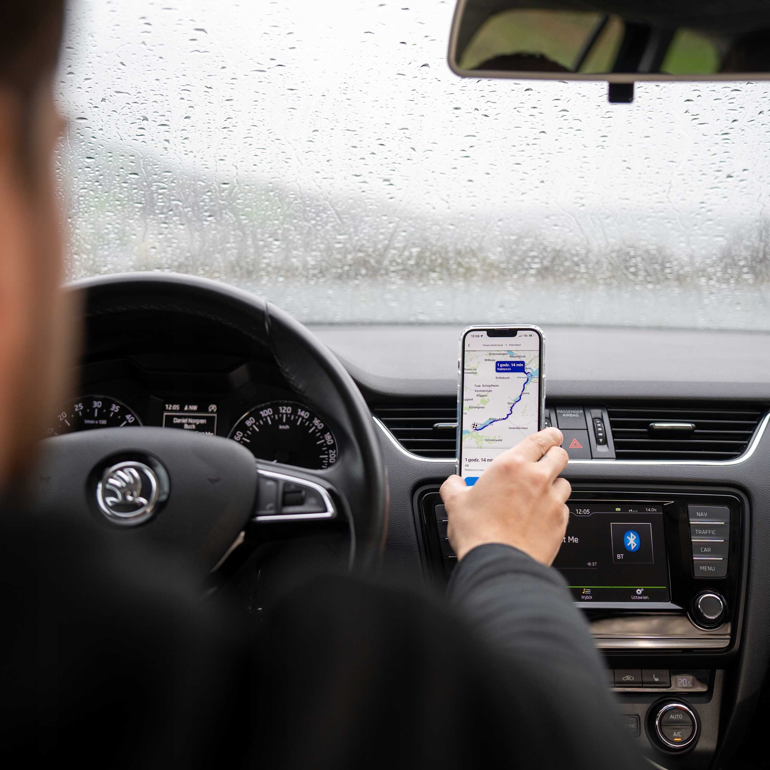 Osoba w samochodzie używa telefonu z aplikacją nawigacyjną. Widoczne krople deszczu na przedniej szybie. Widoczne wnętrze samochodu.