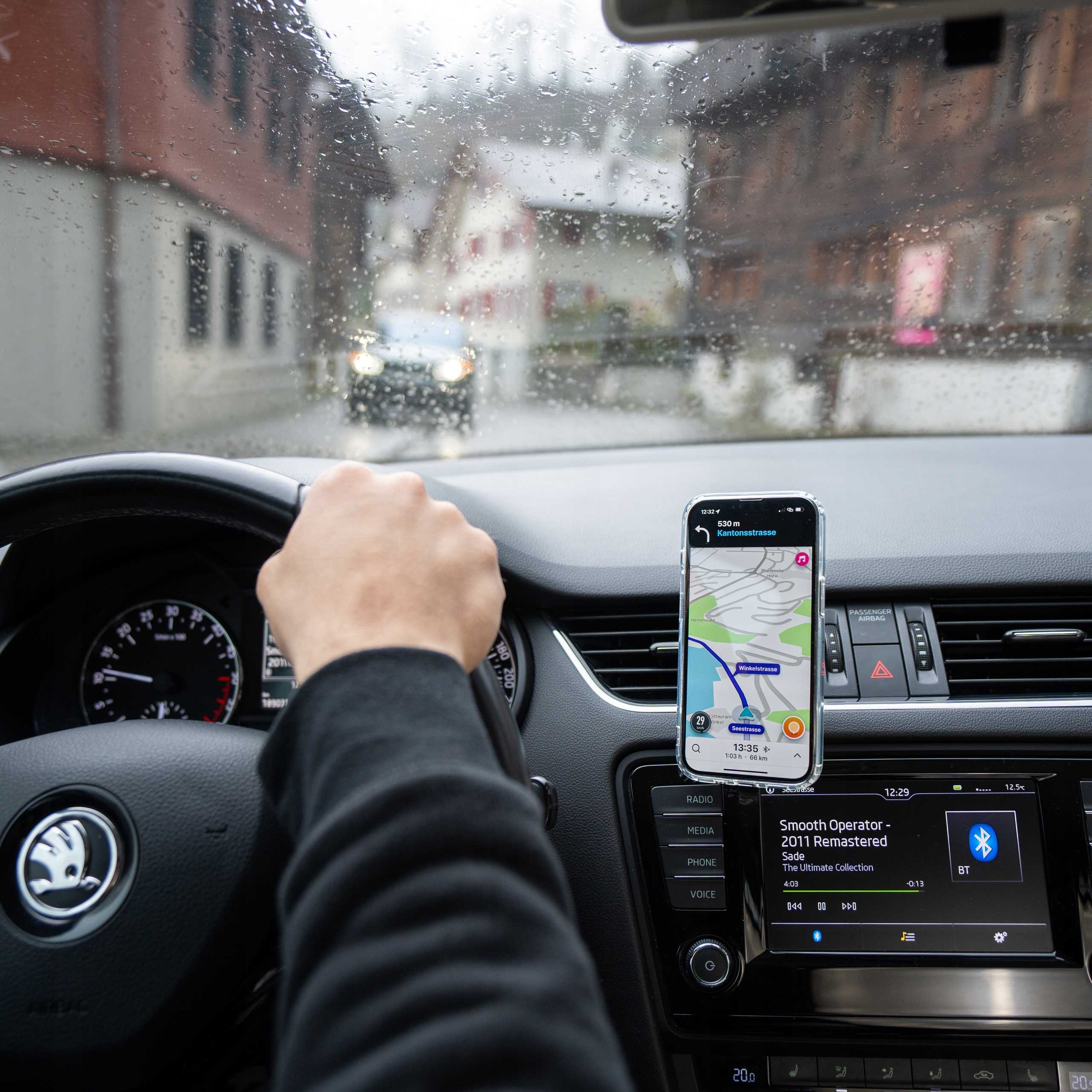 Wewnątrz samochodu, ręka na kierownicy. Telefon wyświetla mapę. Scena oglądana przez mokrą przednią szybę.