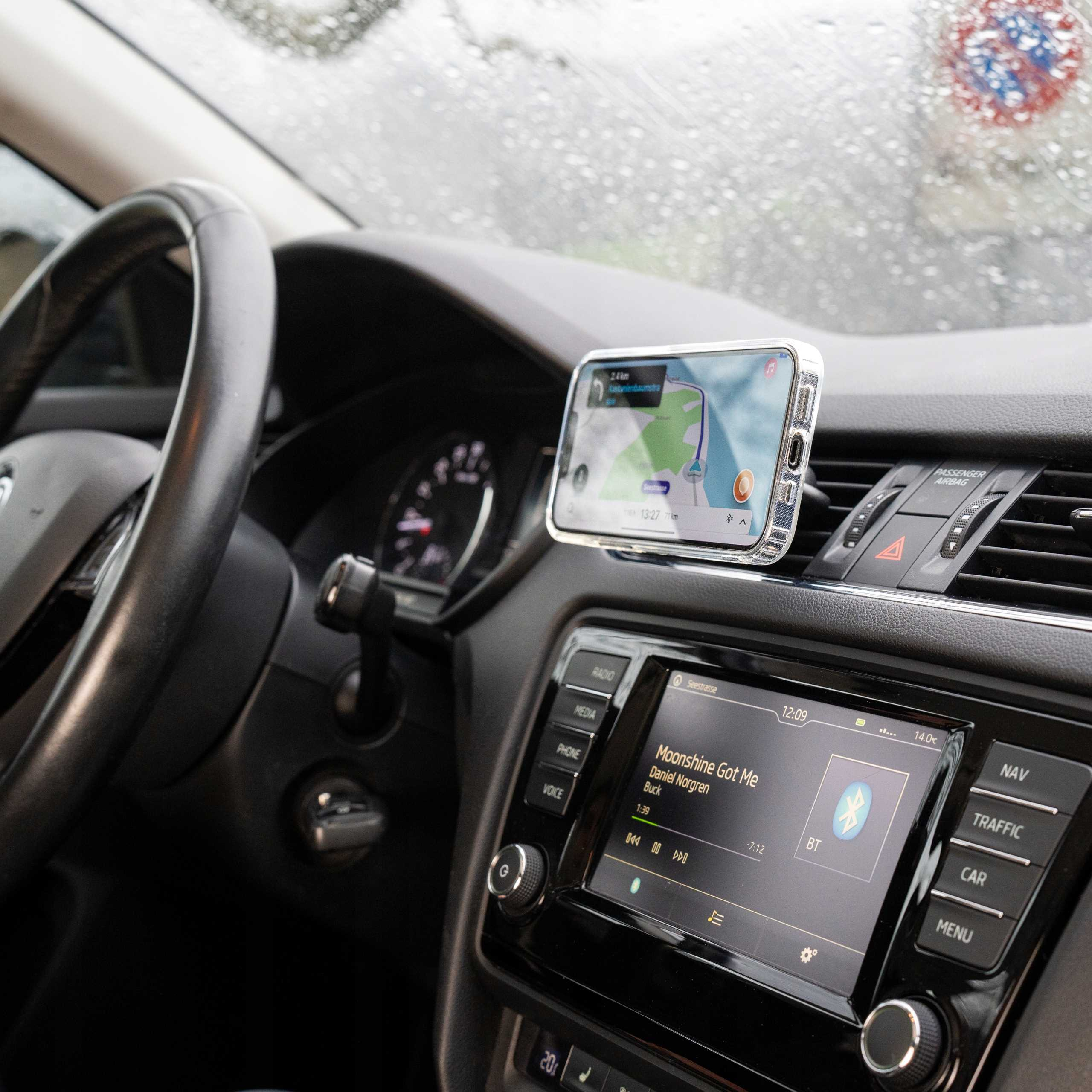 Wnętrze samochodu z kierownicą, telefonem z mapą, radiem i przednią szybą z kroplami deszczu.