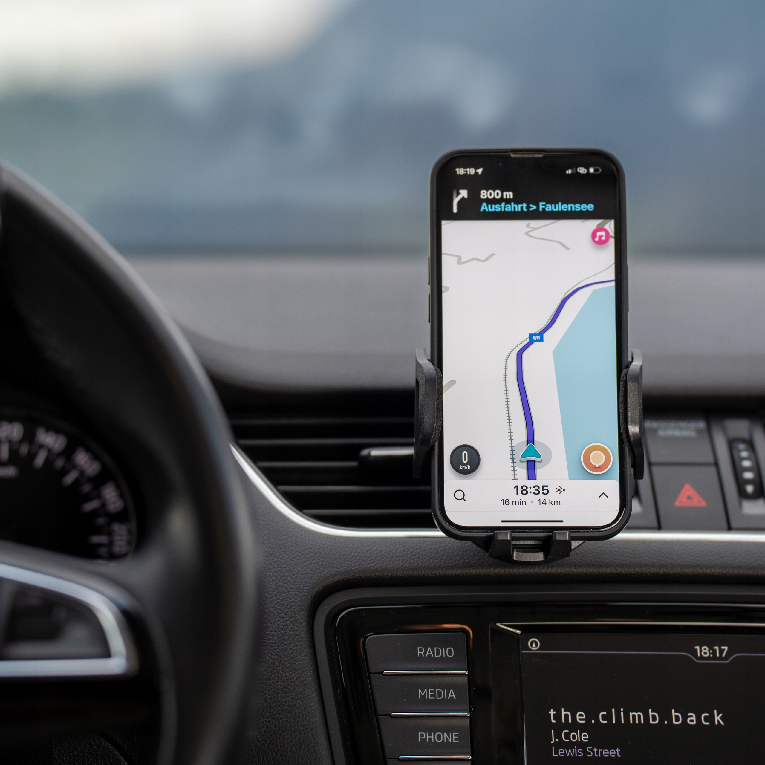 Smartfon w uchwycie samochodowym wyświetla aplikację nawigacyjną. Widoczne jest wnętrze samochodu, w tym deska rozdzielcza.