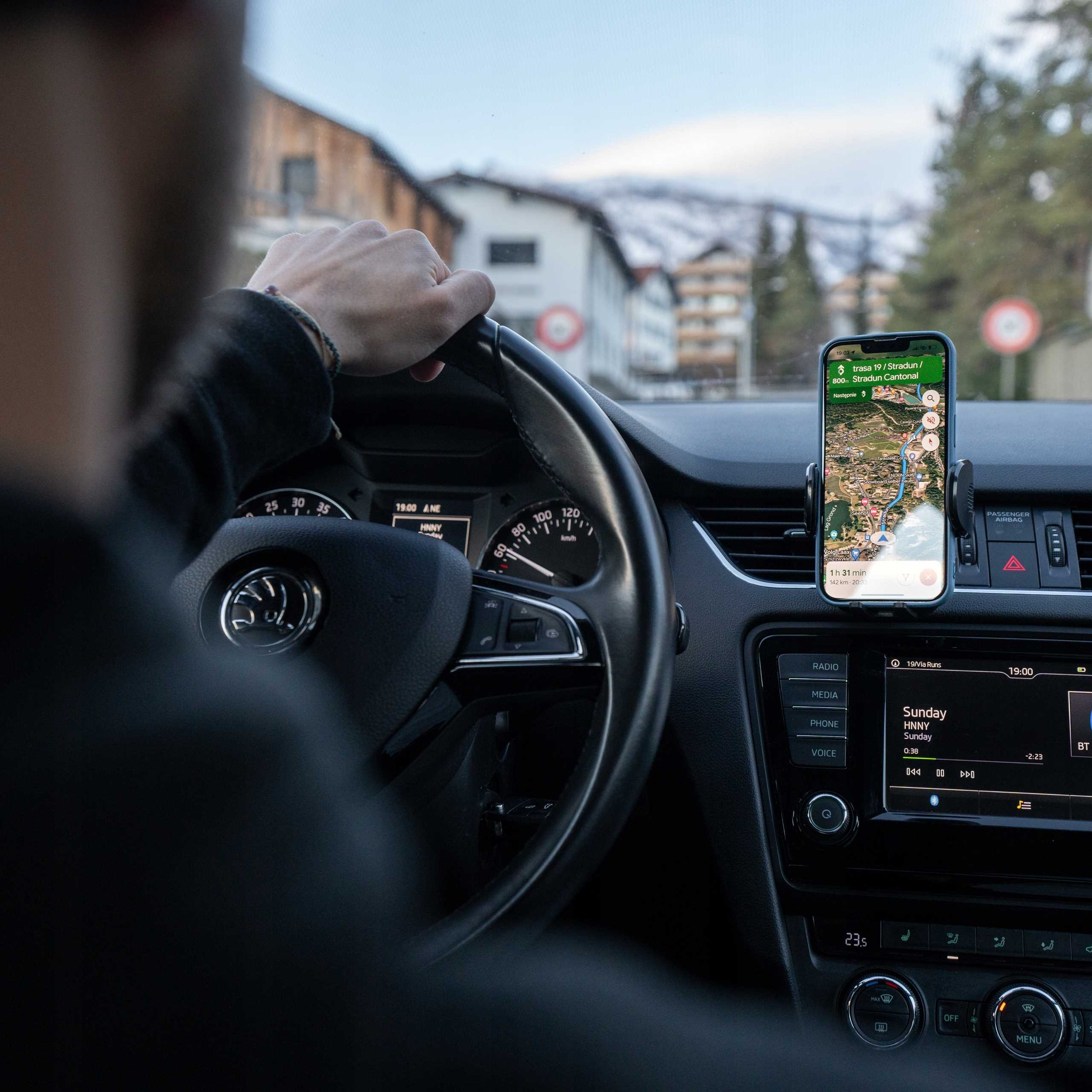 Osoba prowadzi samochód, korzystając z GPS w telefonie zamontowanym na desce rozdzielczej. Widoczne budynki i zaśnieżone wzgórza.