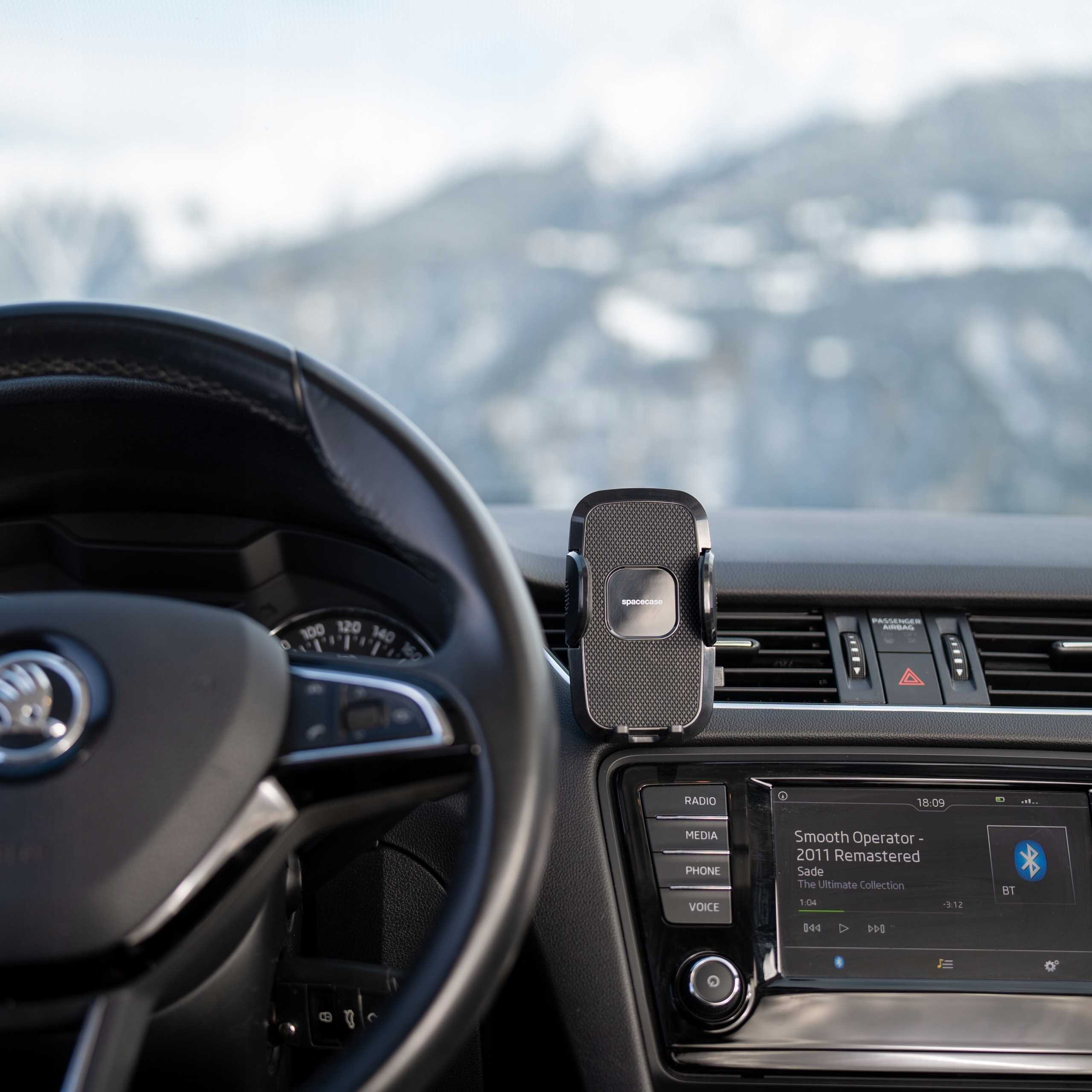 Wnętrze samochodu z uchwytem na telefon, kierownicą i deską rozdzielczą z muzyką i czasem, z rozmytym tłem górskim.