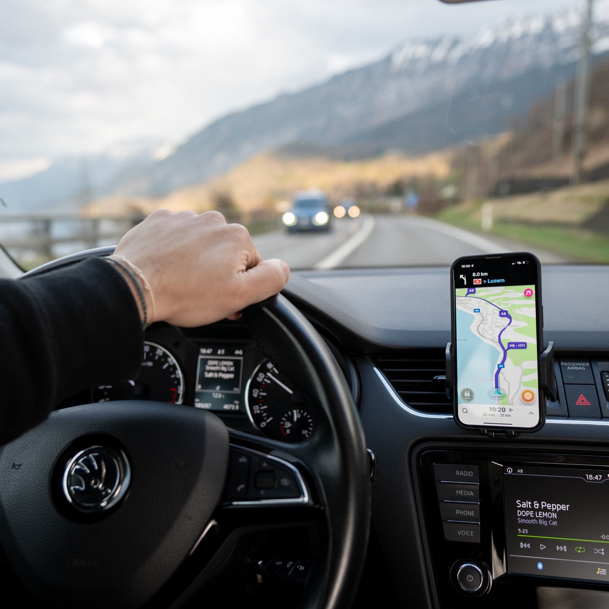 Widok z perspektywy kierowcy w samochodzie. Telefon wyświetla mapę. Widoczne góry. Ręka na kierownicy.