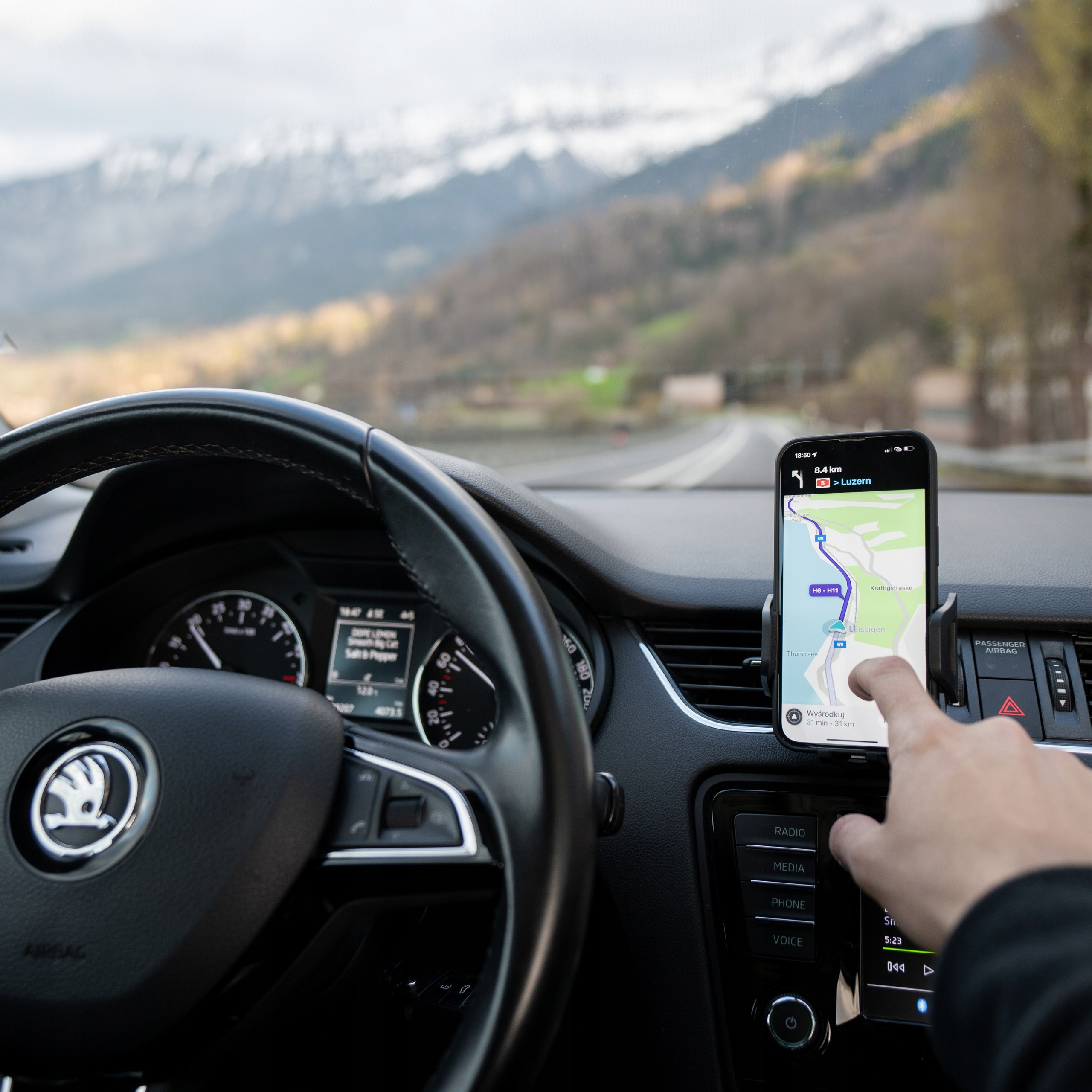 W samochodzie ręka dotyka telefonu z mapą nawigacji. W tle widoczne są góry. Deska rozdzielcza samochodu jest ostra.